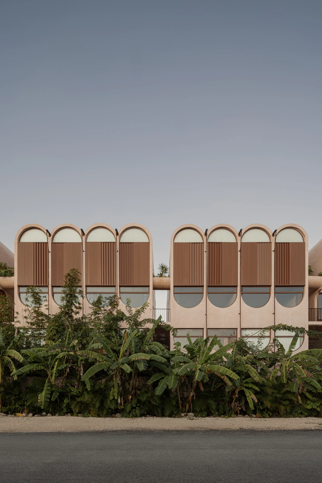 Babel by V Taller, photographed by Funciono — The symmetrical street elevation, arched bays fitted with brown timber louvred shutters over oval windows, screened by a stand of banana palms.