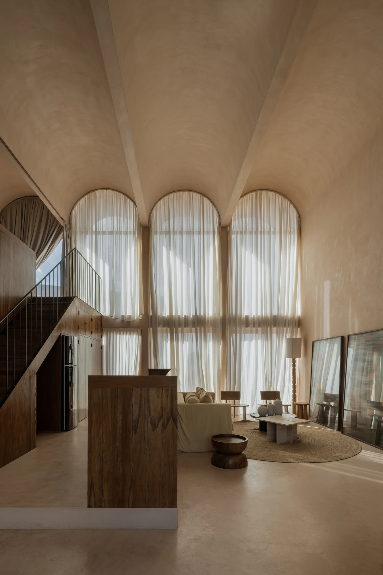 Babel by V Taller, photographed by Funciono — A double-height room under vaults in pale plaster, sheer linen curtains covering full arched windows, a timber counter and a steel-railed stair to one side.
