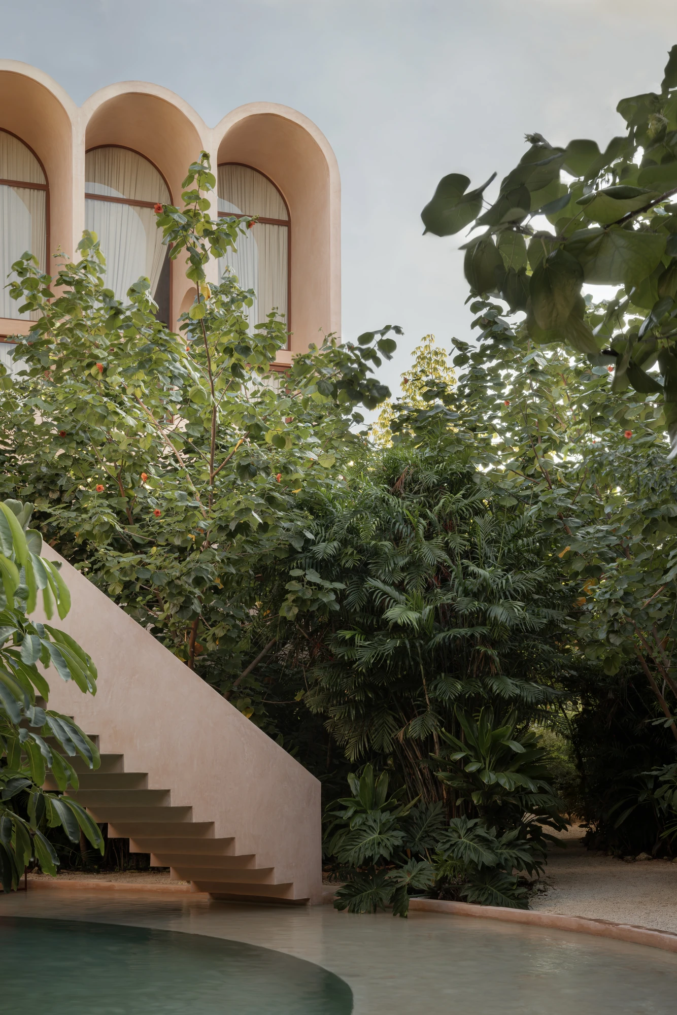 Babel by V Taller, photographed by Funciono — A cantilevered plaster stair with open treads rises beside the pool through hibiscus and palm, the arched upper storey visible above the planting.