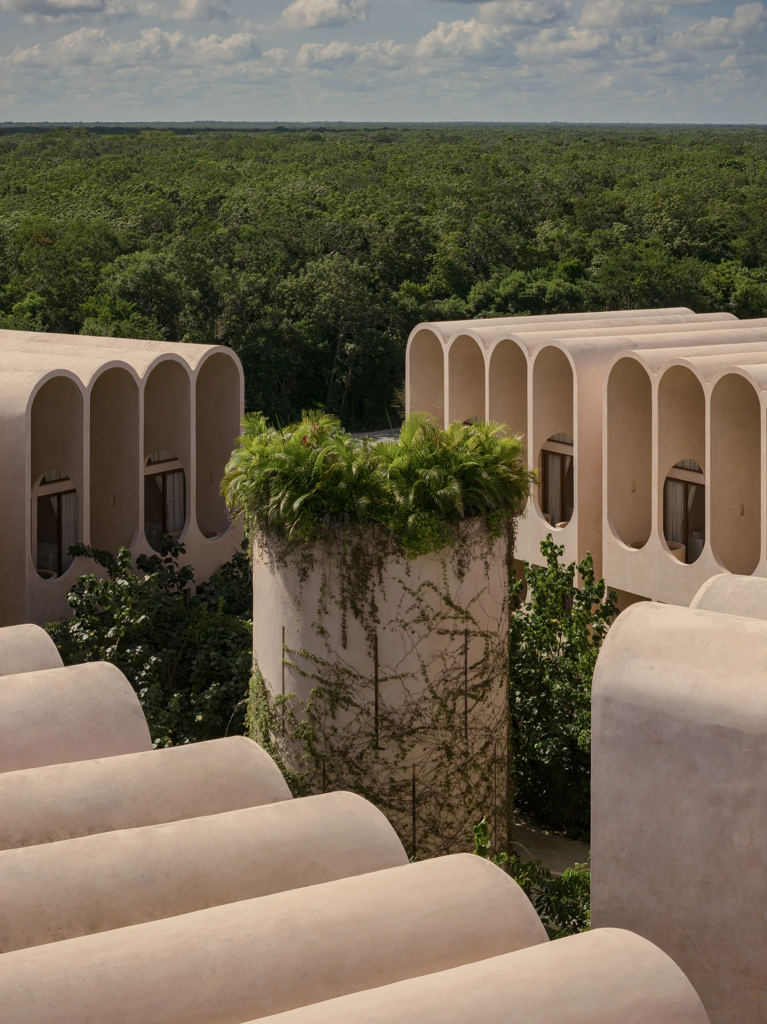 Babel by V Taller, photographed by Funciono — Rows of rounded vault roofs step toward an unbroken jungle horizon, a cylindrical tower rising between them with ferns and palms planted on its crown.