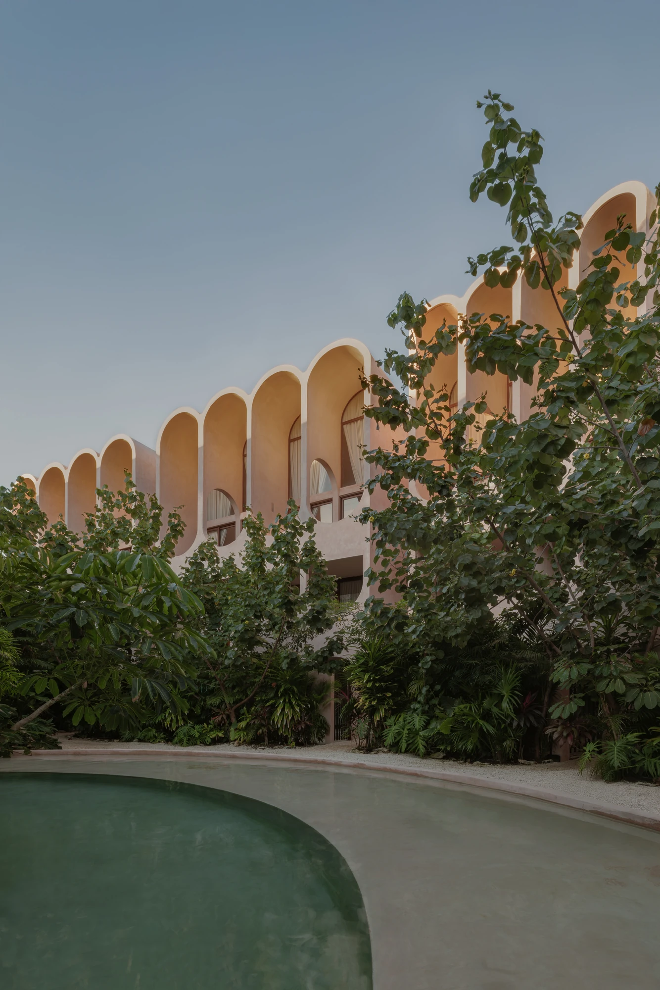 Babel by V Taller, photographed by Funciono — The arcaded wing at dusk above a bank of sea grape and palms, its arches glowing warm, the green pool sweeping across the foreground.
