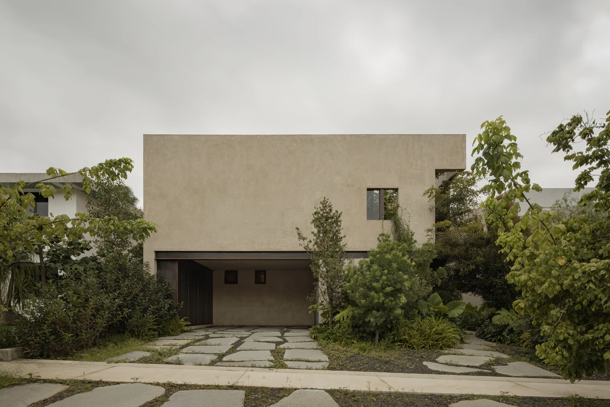 Casa Azaleas by V Taller, photographed by Funciono — The street elevation, a plain cement plaster box above a recessed dark timber gate, approached across irregular stone slabs seamed with low growth.