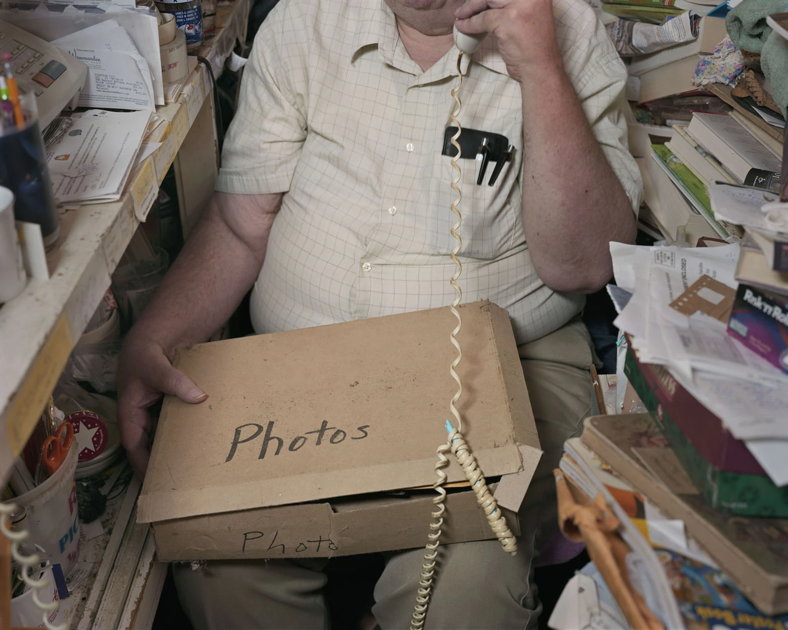 A Pound of Pictures by Alec Soth, photographed by Alec Soth — A man in a checked shirt takes a call on a corded telephone with a cardboard box marked Photos across his knees, from A Pound of Pictures by Alec Soth.