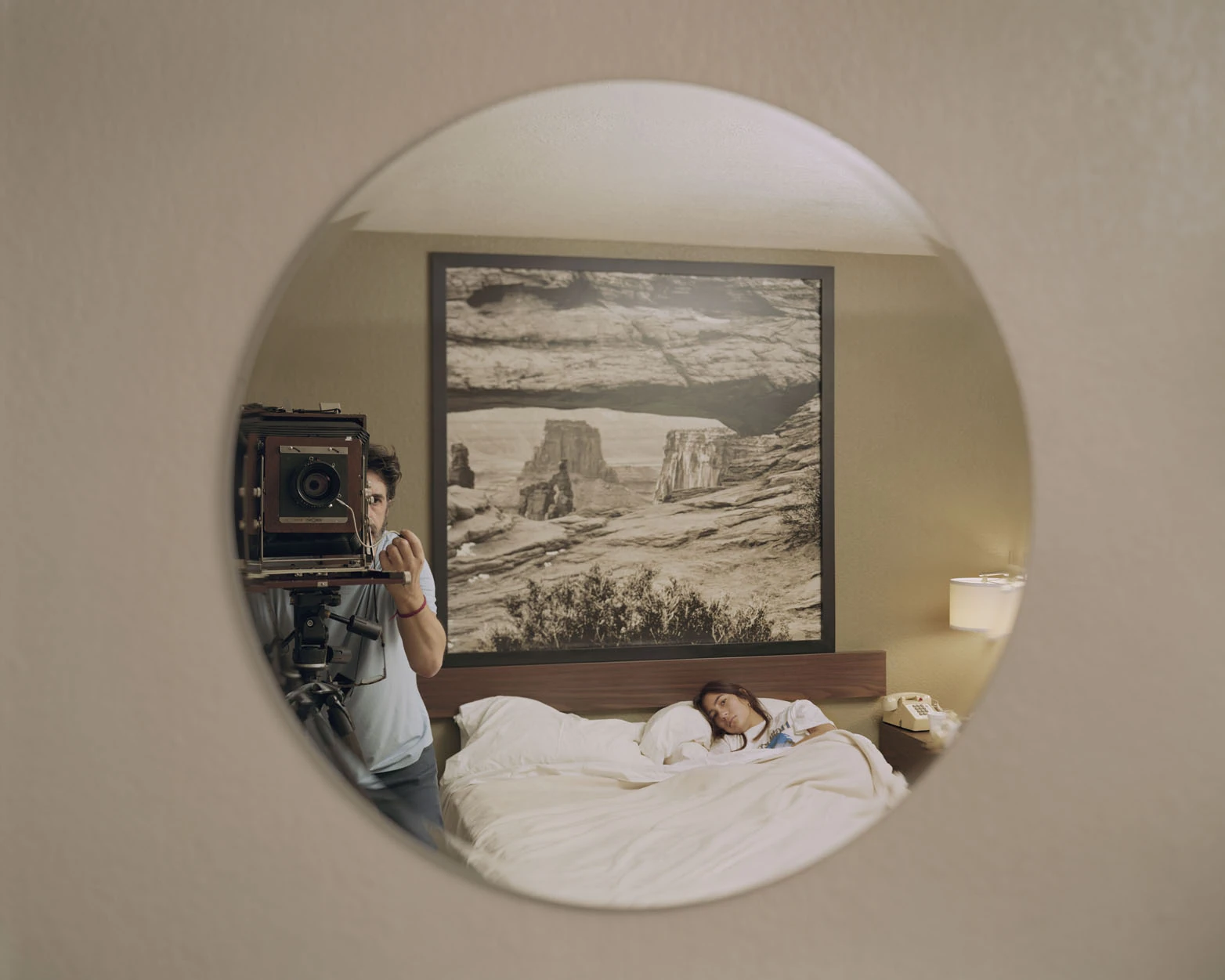 A Pound of Pictures by Alec Soth, photographed by Alec Soth — A round mirror on a motel wall reflects Alec Soth behind a large-format camera, a framed canyon print and a woman lying awake in bed, from A Pound of Pictures by Alec Soth.