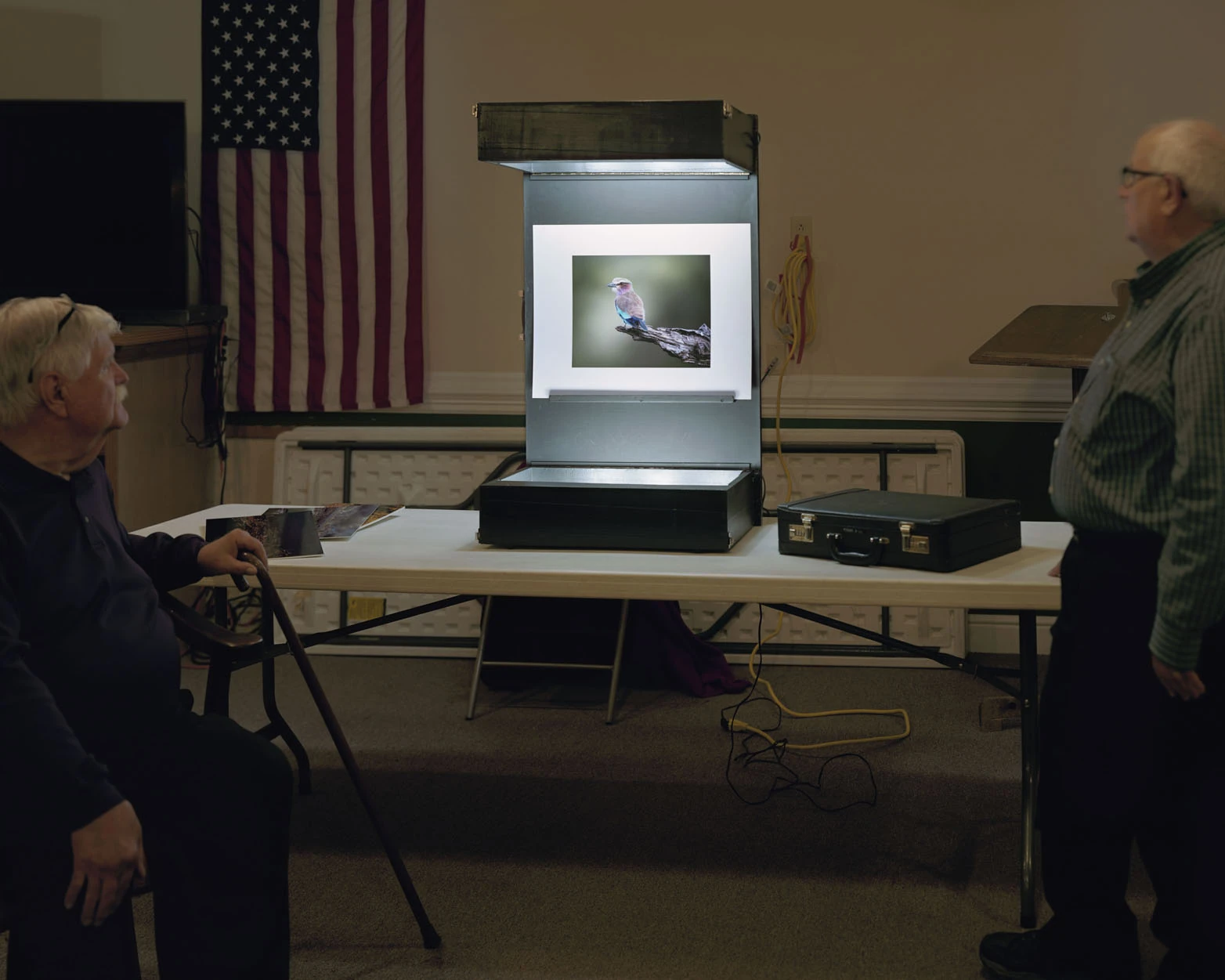 A Pound of Pictures by Alec Soth, photographed by Alec Soth — Two older men flank a lit print-viewing box holding a photograph of a lilac-breasted roller, a US flag hung on the wall behind, from A Pound of Pictures by Alec Soth.