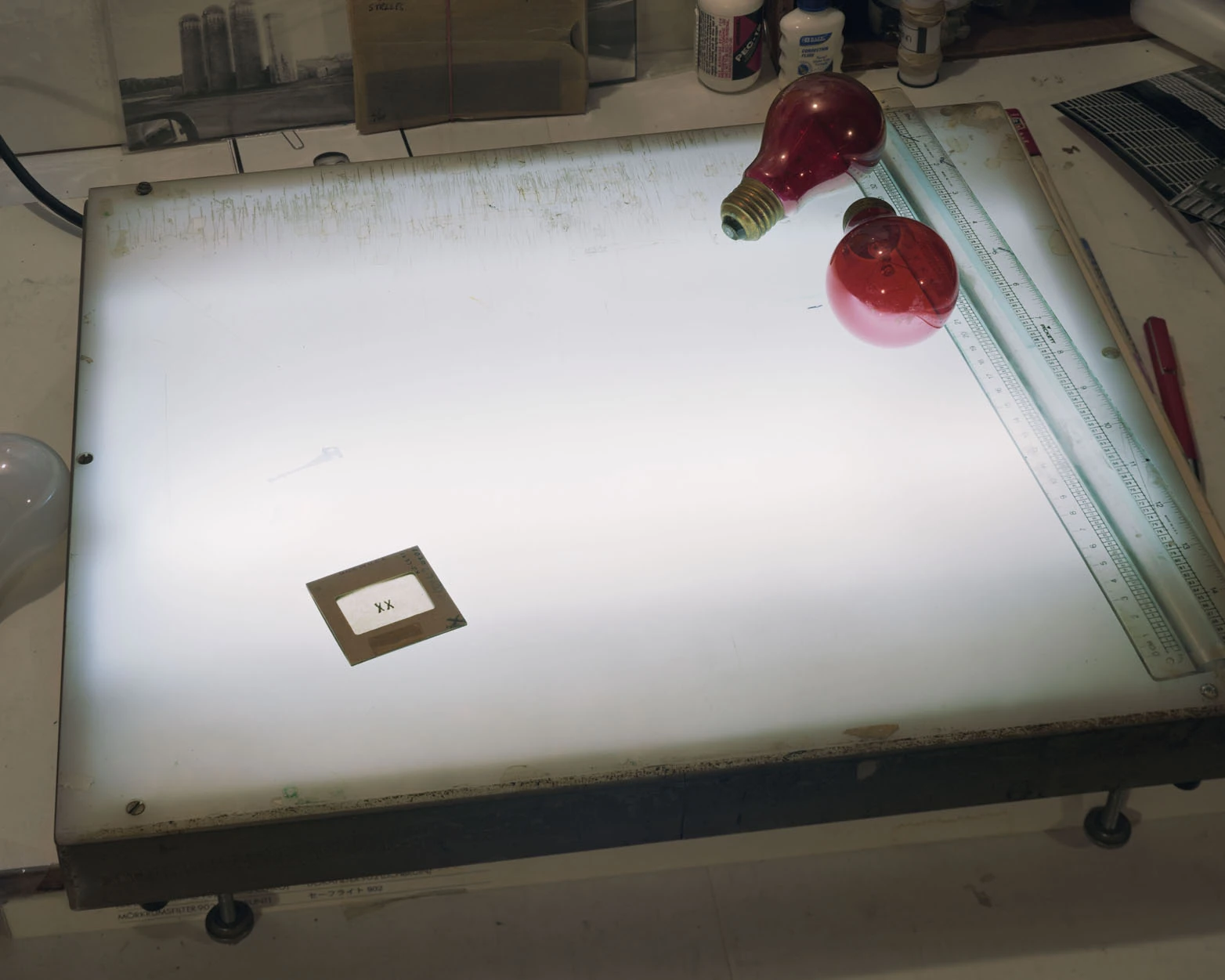 A Pound of Pictures by Alec Soth, photographed by Alec Soth — A single mounted slide sits on an illuminated darkroom light table beside two red bulbs and a steel ruler, from A Pound of Pictures by Alec Soth.