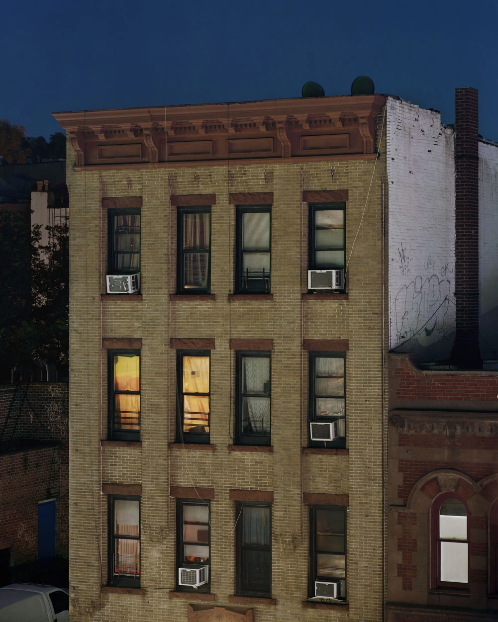 A Pound of Pictures by Alec Soth, photographed by Alec Soth — A four-storey brick apartment block at dusk, two curtained windows burning orange, window units bolted below the sills, from A Pound of Pictures by Alec Soth.