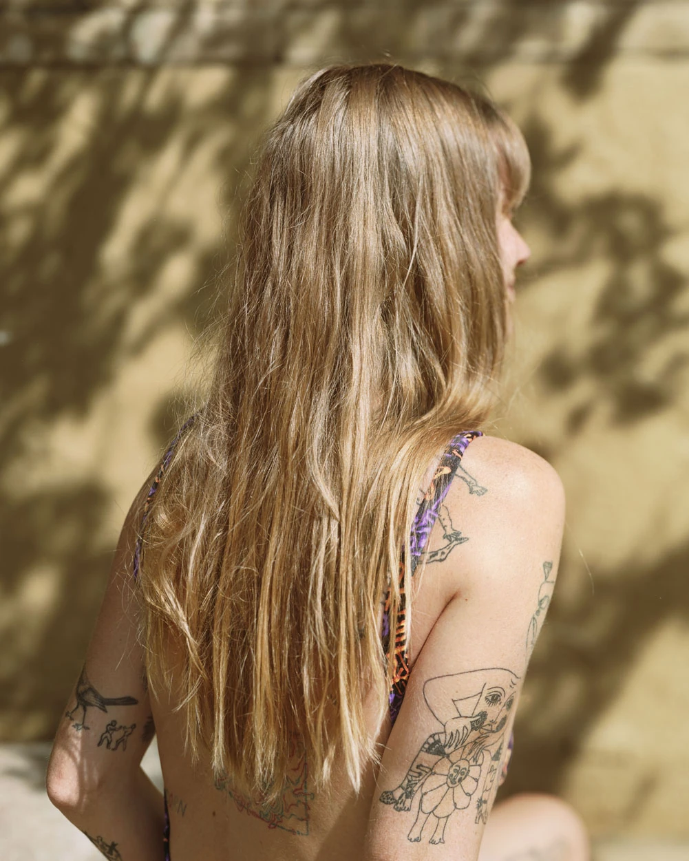 A Pound of Pictures by Alec Soth, photographed by Alec Soth — A woman seen from behind, long blonde hair and tattooed shoulders against an ochre wall in leaf shadow, from A Pound of Pictures by Alec Soth.