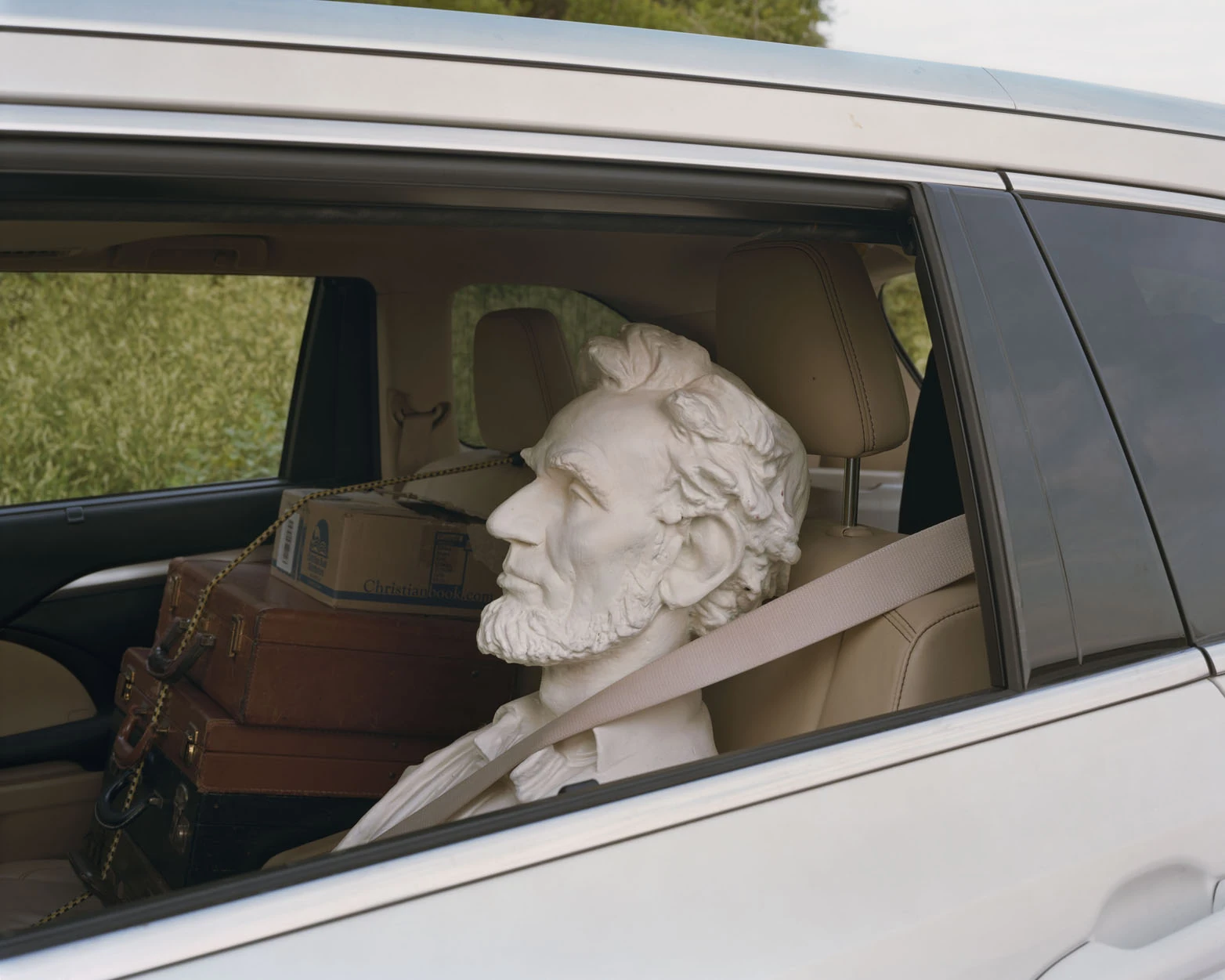 A Pound of Pictures by Alec Soth, photographed by Alec Soth — A white plaster bust of Abraham Lincoln belted into a car's back seat beside leather suitcases and a cardboard box, from A Pound of Pictures by Alec Soth.