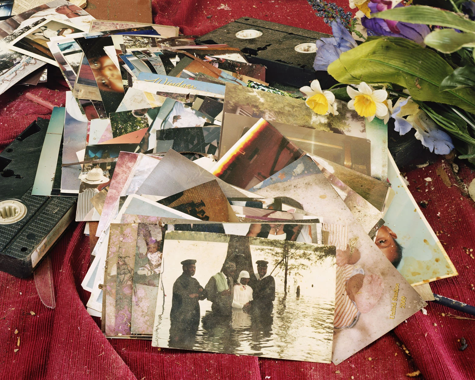 A Pound of Pictures by Alec Soth, photographed by Alec Soth — Water-stained snapshots heaped on red corduroy among VHS cassettes and artificial flowers, a baptism in floodwater near the top, from A Pound of Pictures by Alec Soth.
