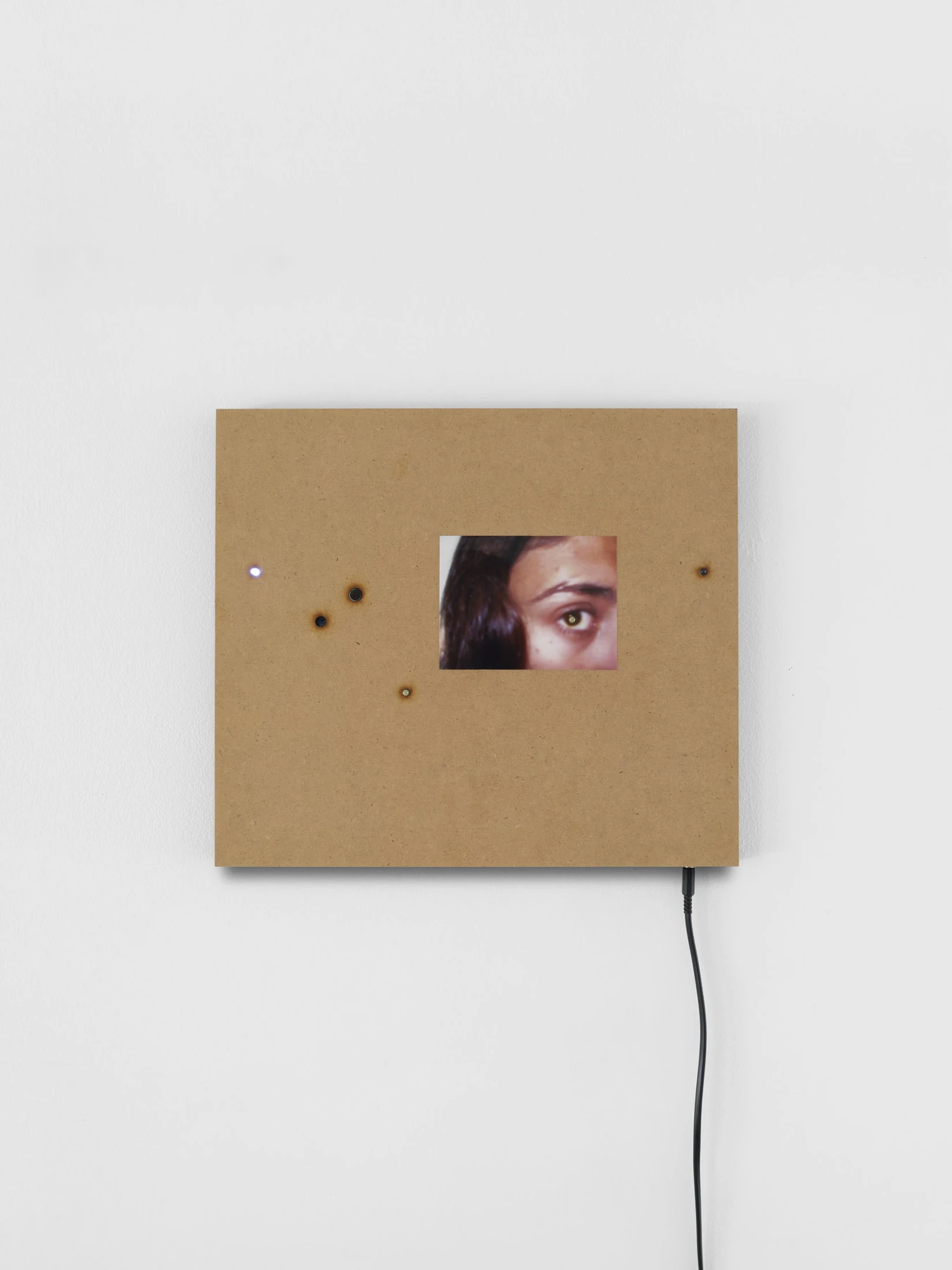 Apophenia by Galerie Molitor, photographed by Marjorie Brunet-Plaza — A raw MDF square with a small screen showing an eye and brow, brass studs and a pinpoint LED set into the board.