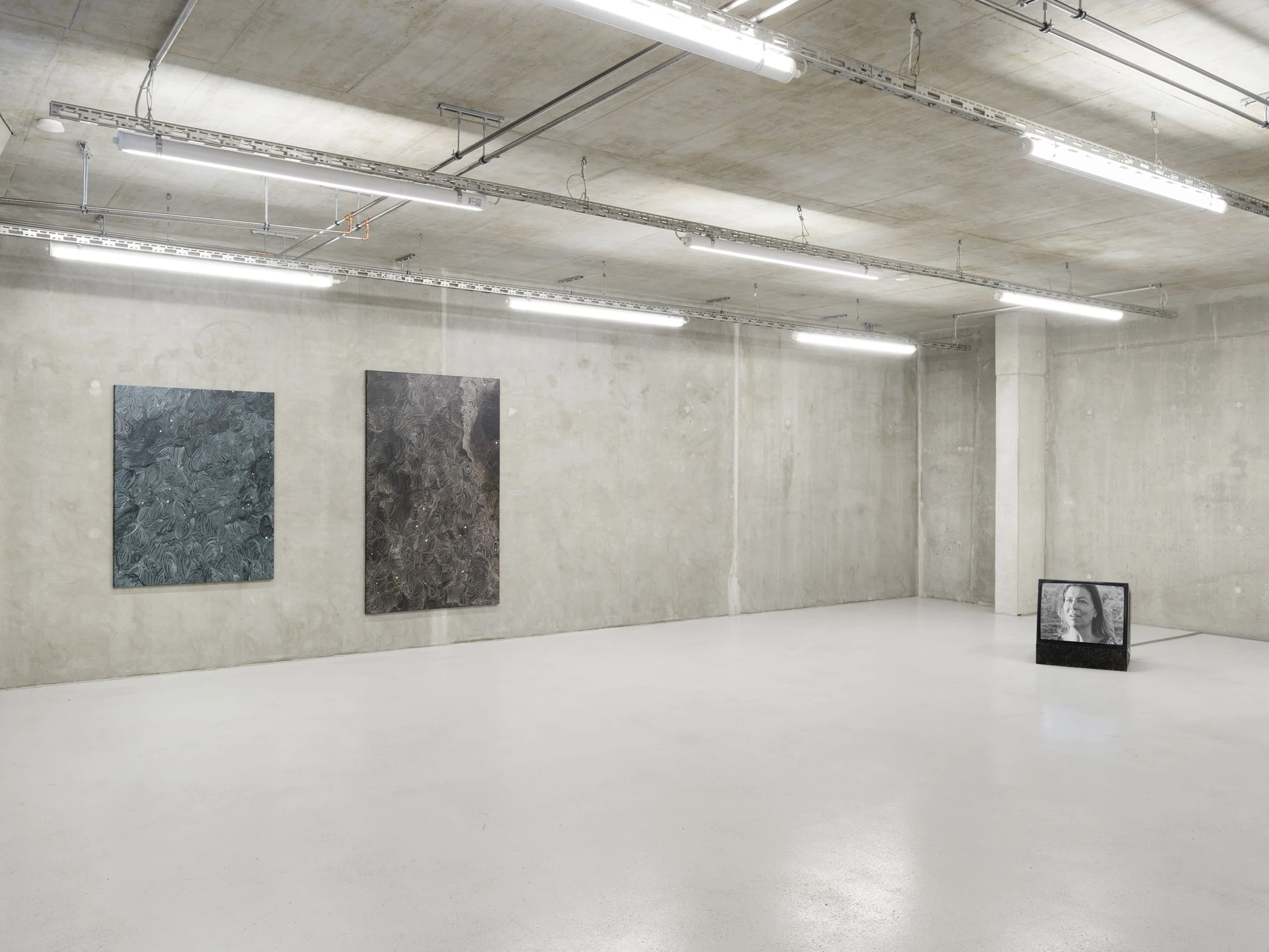 Apophenia by Galerie Molitor, photographed by Marjorie Brunet-Plaza — The same concrete room, the corner monitor now showing a woman's face in black and white.