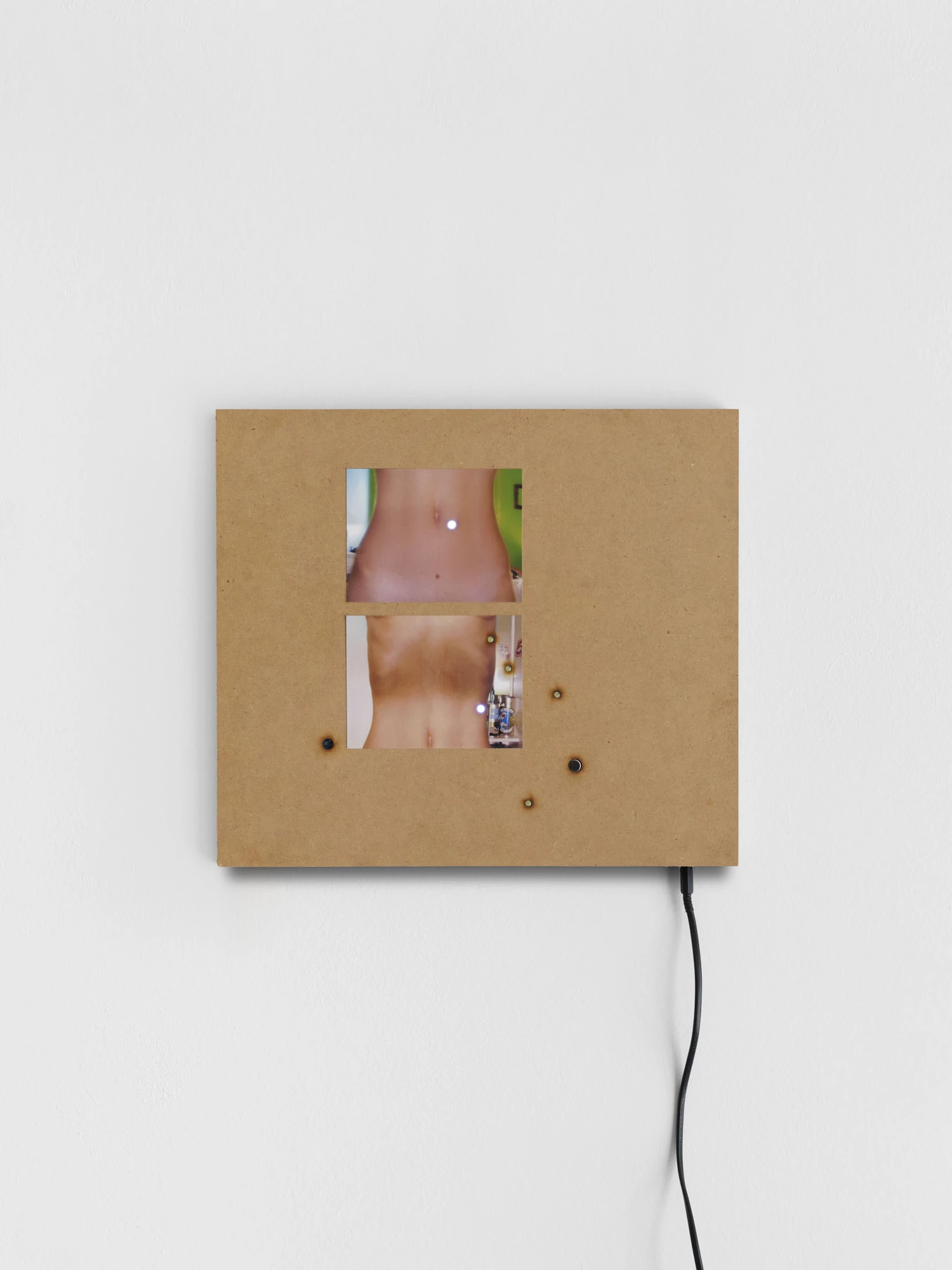 Apophenia by Galerie Molitor, photographed by Marjorie Brunet-Plaza — A raw MDF square with two stacked screens showing a bare torso and navel, studs and lit LEDs across the board.