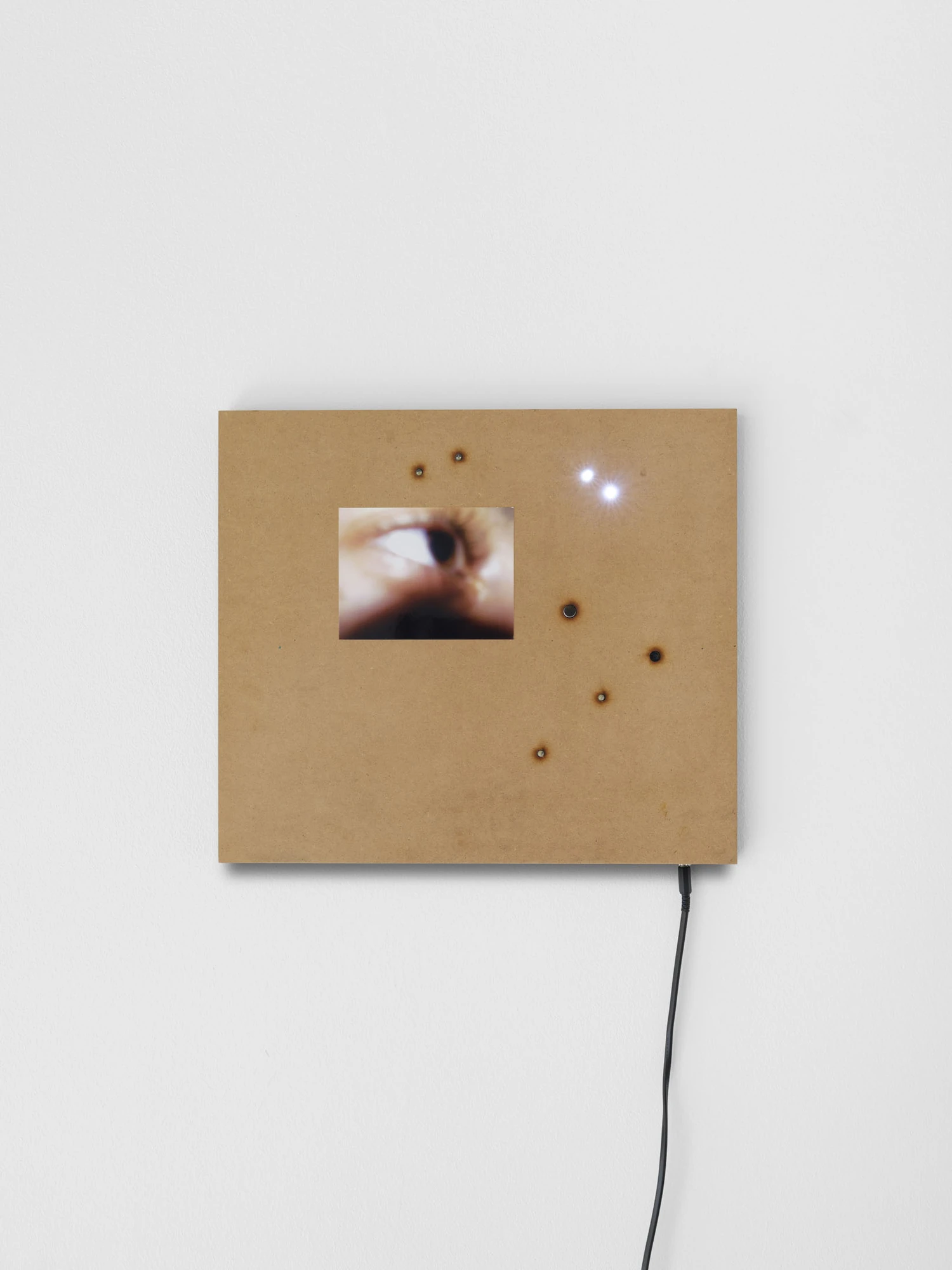 Apophenia by Galerie Molitor, photographed by Marjorie Brunet-Plaza — A raw MDF square with a screen showing an eye at extreme magnification, two LEDs burning at the upper right.