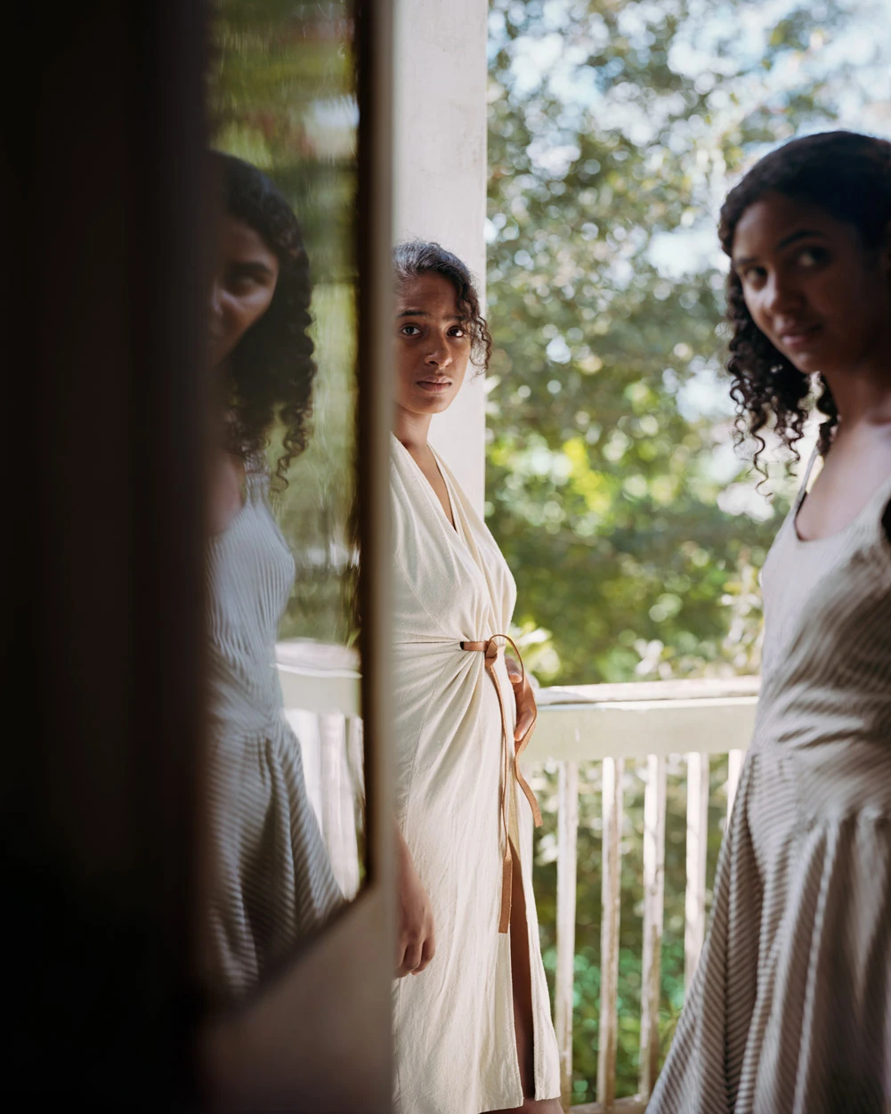 I Know How Furiously Your Heart Is Beating by Alec Soth, photographed by Alec Soth — A woman in a cream wrap dress on a shaded porch, doubled by a mirror leaning at the edge of the frame, from I Know How Furiously Your Heart Is Beating by Alec Soth.