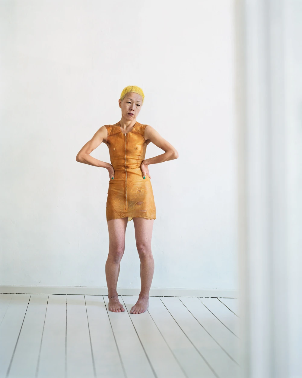 I Know How Furiously Your Heart Is Beating by Alec Soth, photographed by Alec Soth — A woman with bleached yellow hair stands barefoot on white floorboards in a rust-orange latex dress, hands on her hips, from I Know How Furiously Your Heart Is Beating…