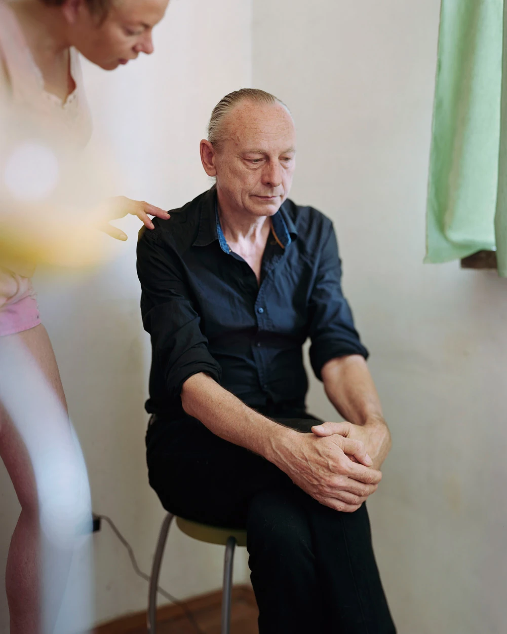 I Know How Furiously Your Heart Is Beating by Alec Soth, photographed by Alec Soth — A man in a black shirt sits on a stool with his hands clasped while a woman's hand rests on his shoulder, from I Know How Furiously Your Heart Is Beating by Alec Soth.