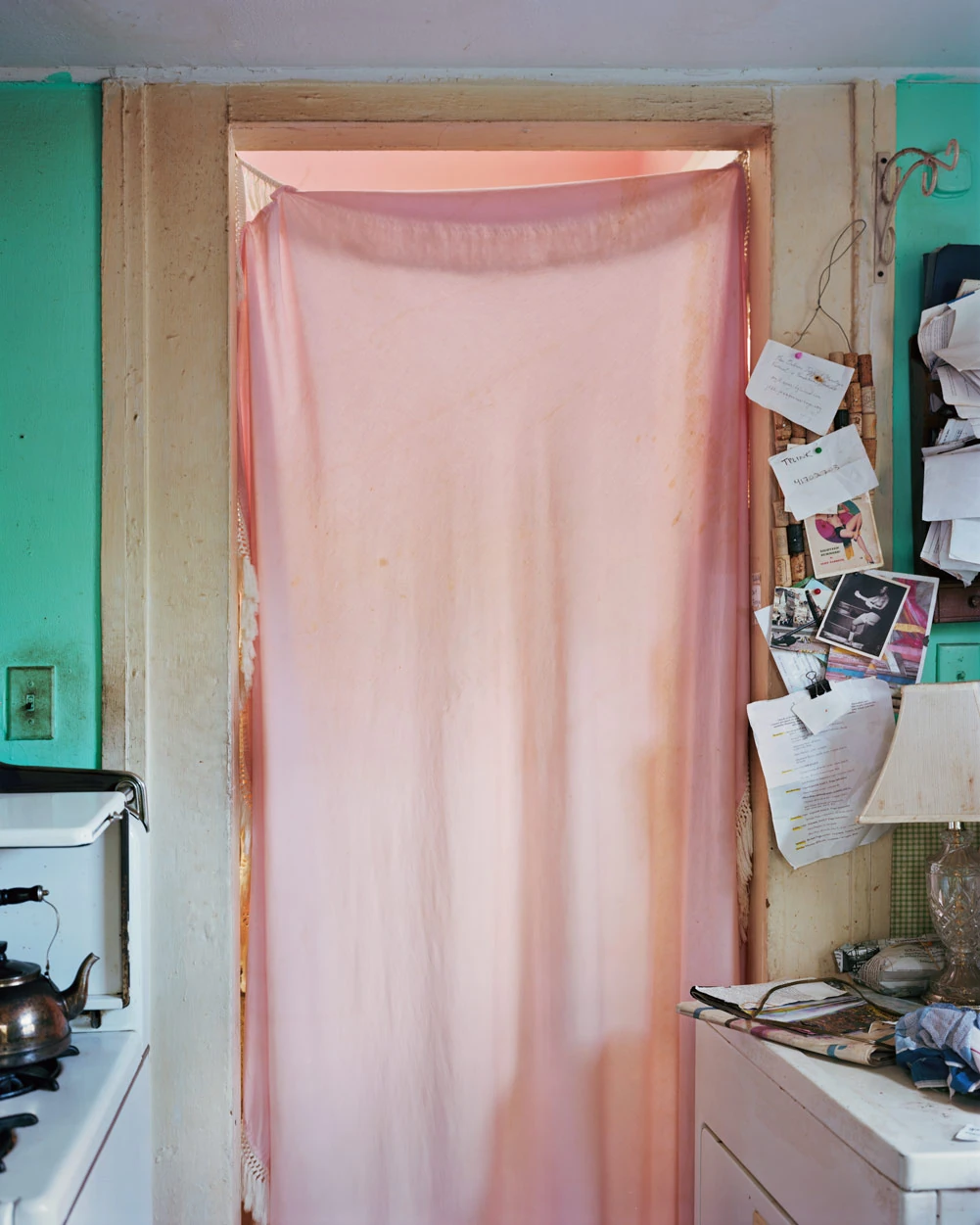 I Know How Furiously Your Heart Is Beating by Alec Soth, photographed by Alec Soth — A pink cloth hung across a kitchen doorway glows from the room behind, notes and clippings pinned to the frame, from I Know How Furiously Your Heart Is Beating by Alec…