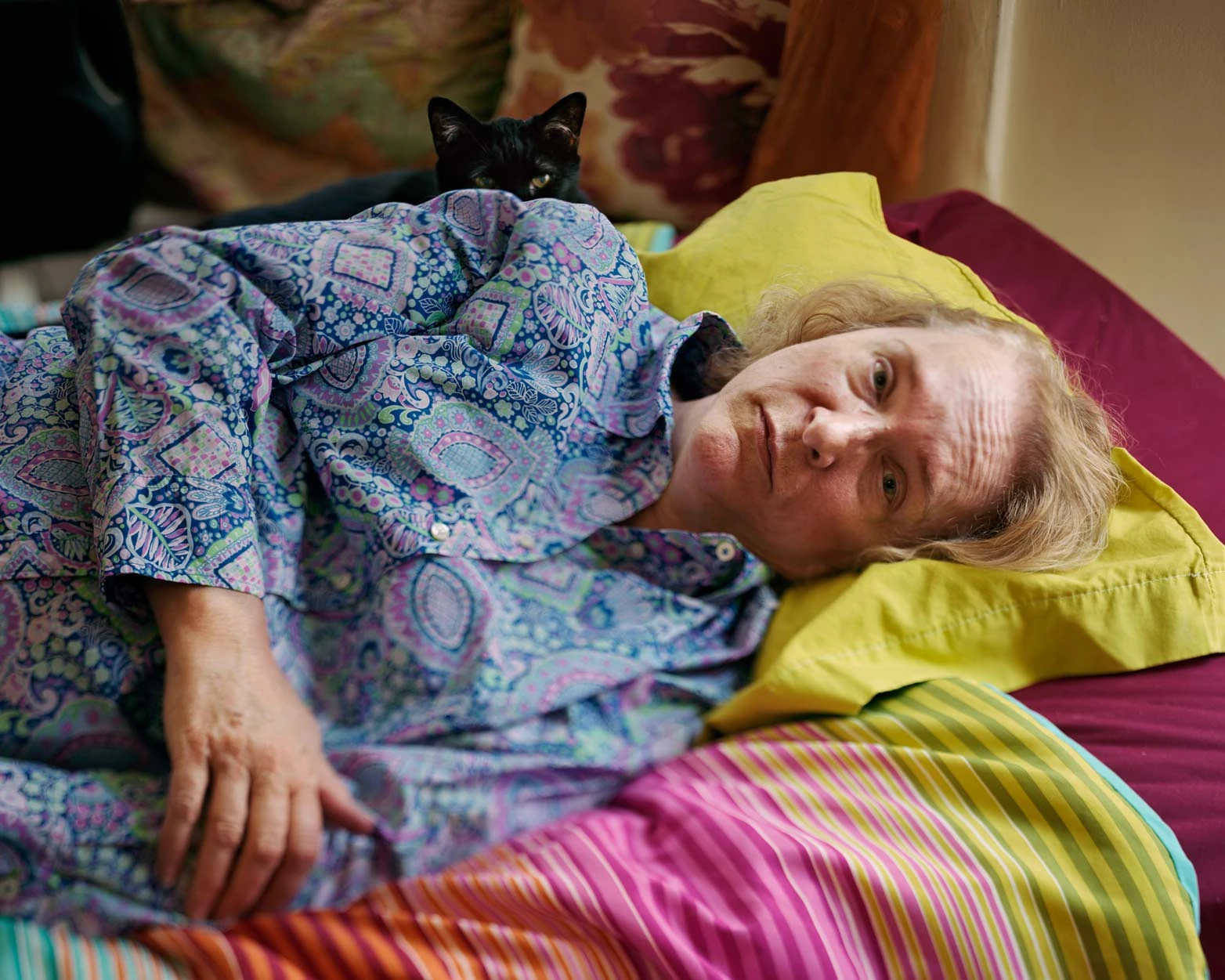 I Know How Furiously Your Heart Is Beating by Alec Soth, photographed by Alec Soth — A woman in a paisley shirt lies on striped and yellow bedding with a black cat pressed against her shoulder, from I Know How Furiously Your Heart Is Beating by Alec Soth.