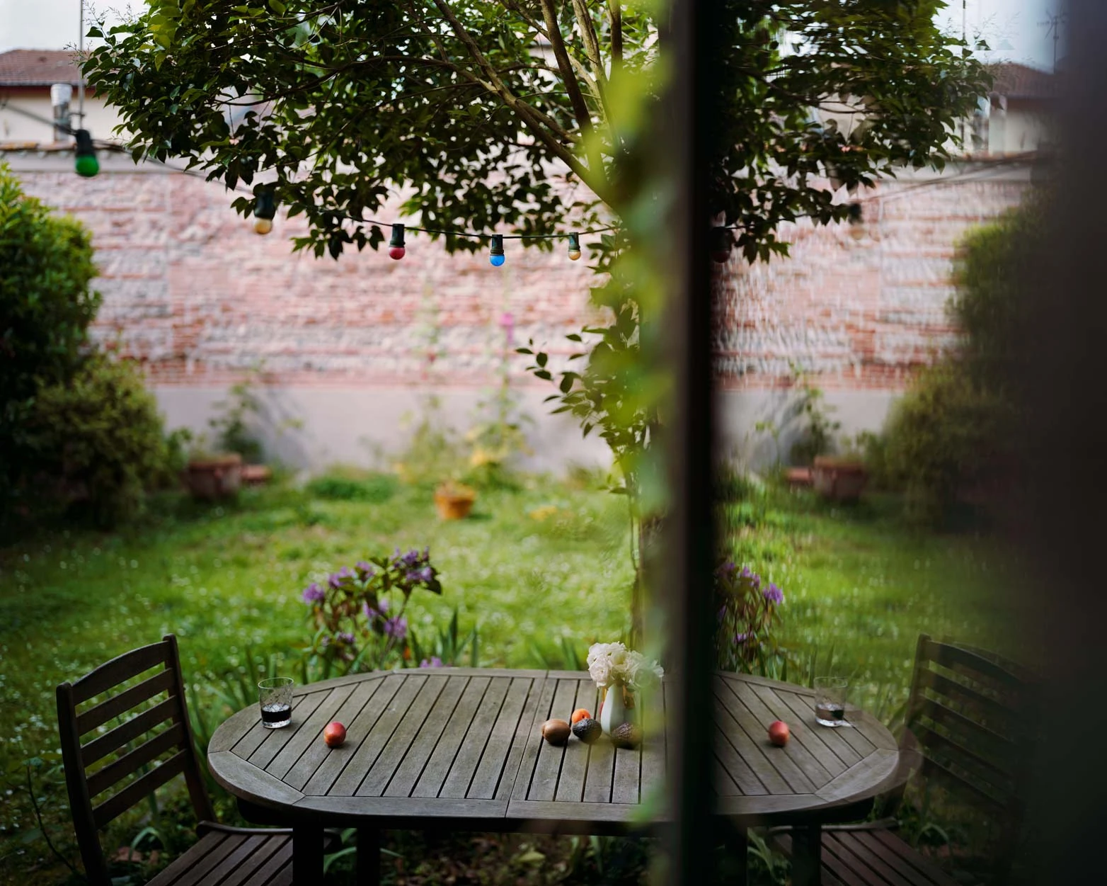 I Know How Furiously Your Heart Is Beating by Alec Soth, photographed by Alec Soth — A garden table set with glasses, fruit and white flowers seen through a doorway, festoon bulbs strung beneath a tree, from I Know How Furiously Your Heart Is Beating by…
