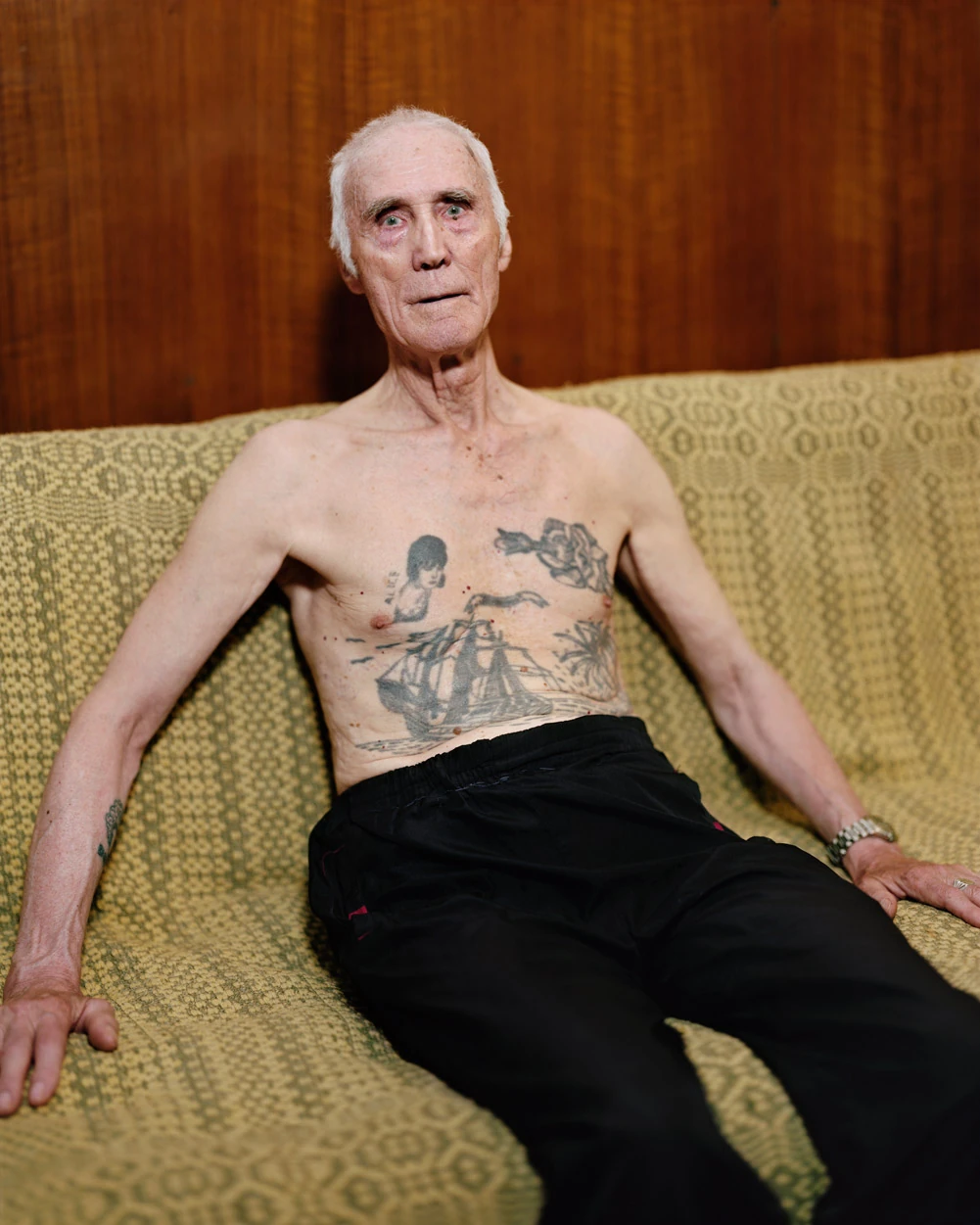 I Know How Furiously Your Heart Is Beating by Alec Soth, photographed by Alec Soth — A shirtless older man reclines on a green woven couch, a full-rigged ship tattooed across his chest, from I Know How Furiously Your Heart Is Beating by Alec Soth.