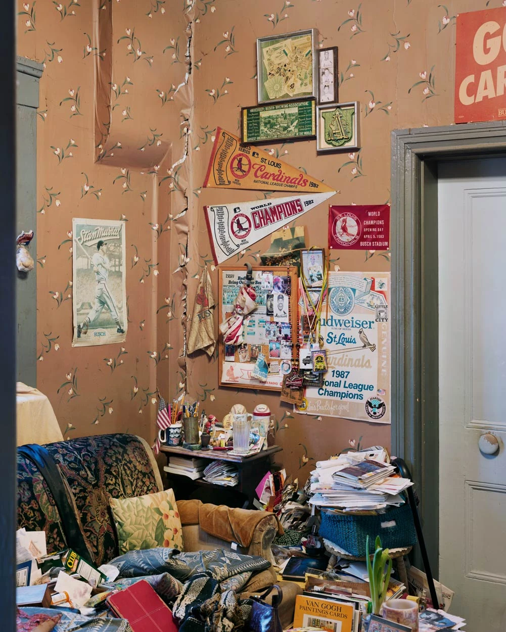 I Know How Furiously Your Heart Is Beating by Alec Soth, photographed by Alec Soth — A corner papered in tulips and hung with St Louis Cardinals pennants above a heaped armchair and stacks of paper, from I Know How Furiously Your Heart Is Beating by…
