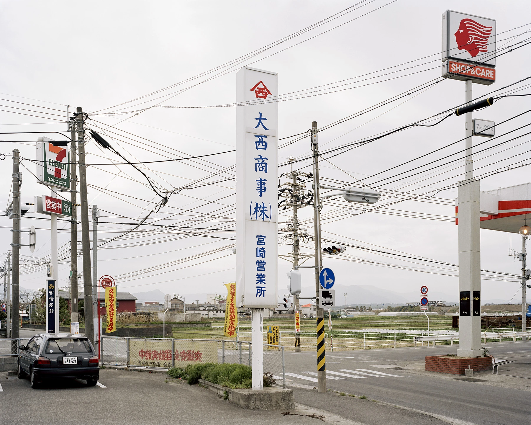 MIYAZAKI by Mark Power, photographed by Mark Power — A rural crossroads dense with signage, a tall white company pylon at the centre, a convenience store sign at left and a petrol station canopy at right, overhead cables crossing a grey sky above rice…