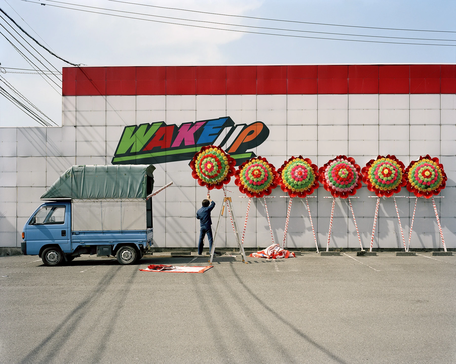MIYAZAKI by Mark Power, photographed by Mark Power — A man on a stepladder adjusting the first of six circular flower wreaths set on tripods along a white panelled wall painted with the word WAKE UP, a small blue truck parked at left.