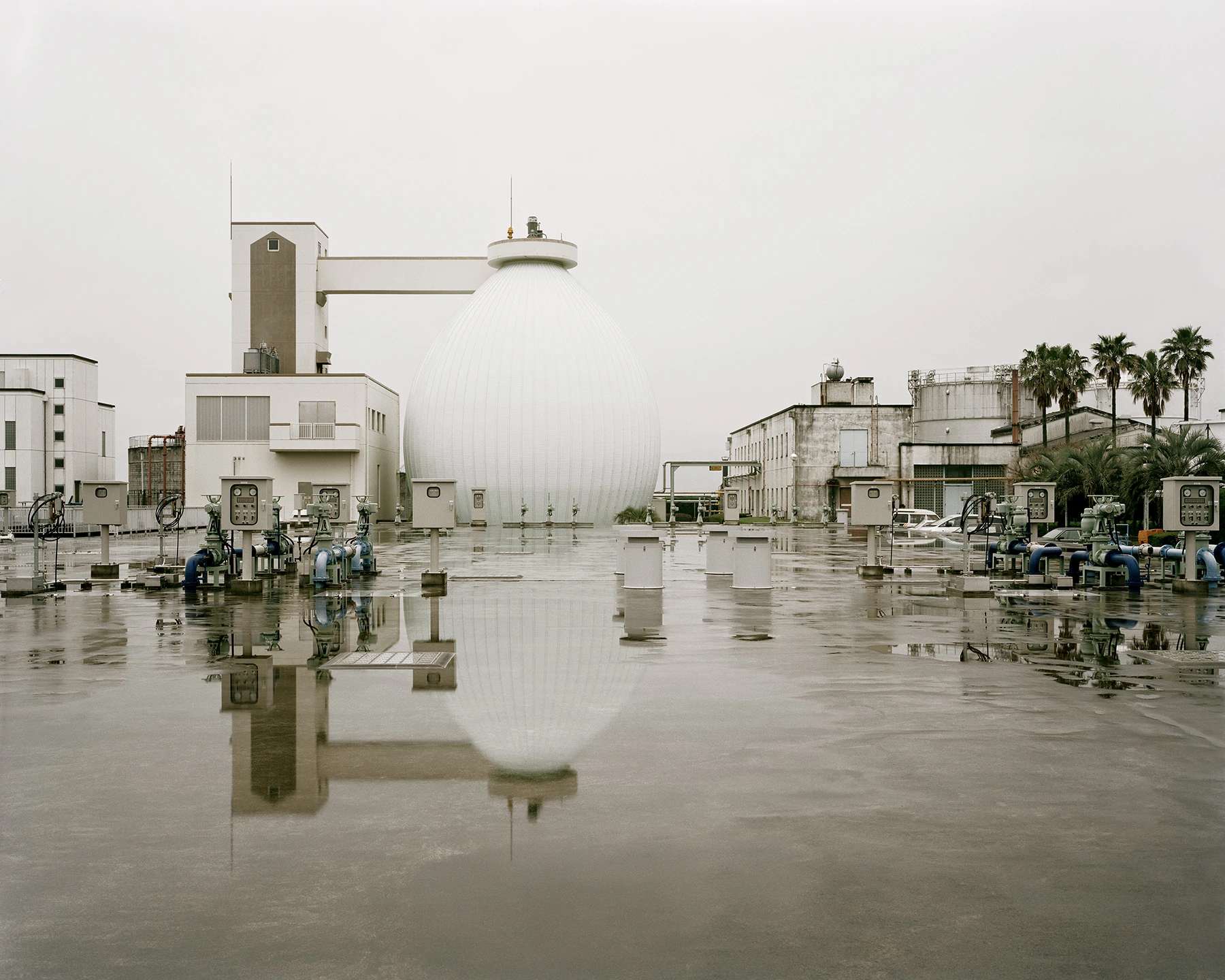 MIYAZAKI by Mark Power, photographed by Mark Power — A white egg-shaped digester tank rising above a treatment plant, its ribbed surface and the surrounding valve gear mirrored in a sheet of standing rainwater across the foreground.