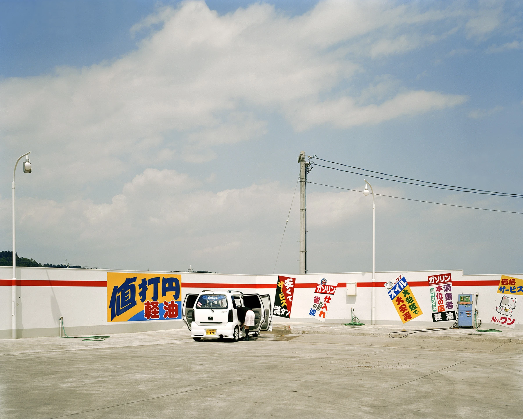 MIYAZAKI by Mark Power, photographed by Mark Power — A white minivan with both doors open on a bare concrete forecourt, a woman leaning into the back, hand-painted price signs ranged along a low white wall behind.