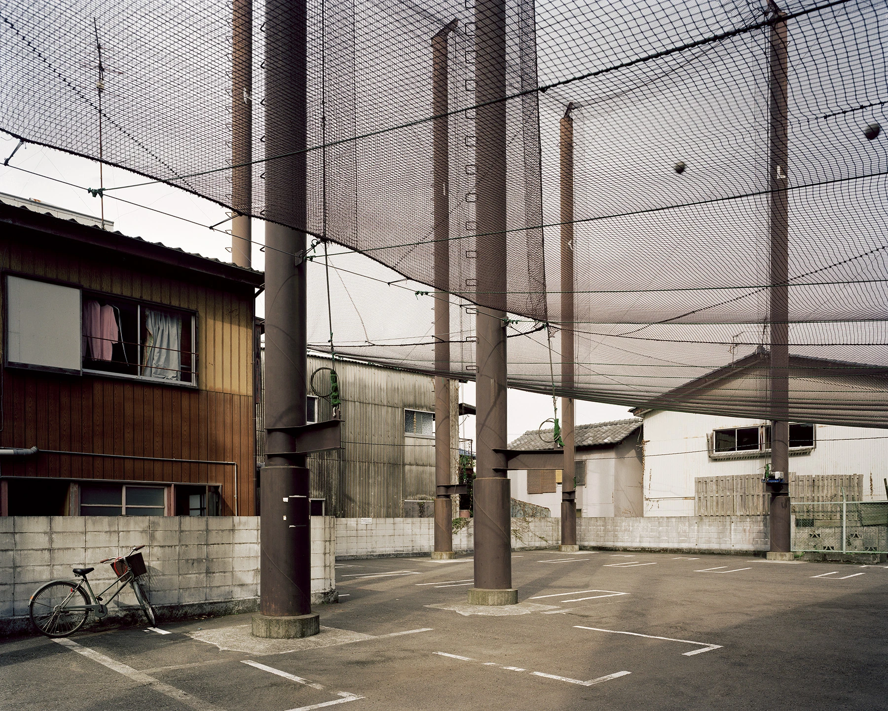 MIYAZAKI by Mark Power, photographed by Mark Power — Rusted steel columns carrying a black batting-cage net above a marked parking lot, a bicycle leaning against a concrete block wall beside a timber-clad house.