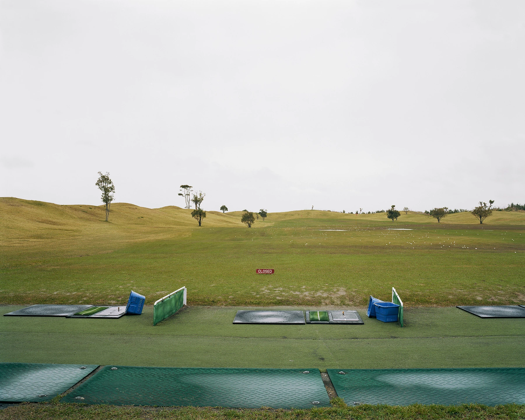 MIYAZAKI by Mark Power, photographed by Mark Power — A driving range with rubber tee mats and blue ball baskets in the foreground, white balls scattered across mown grass, a small red CLOSED sign standing at the centre of the frame.