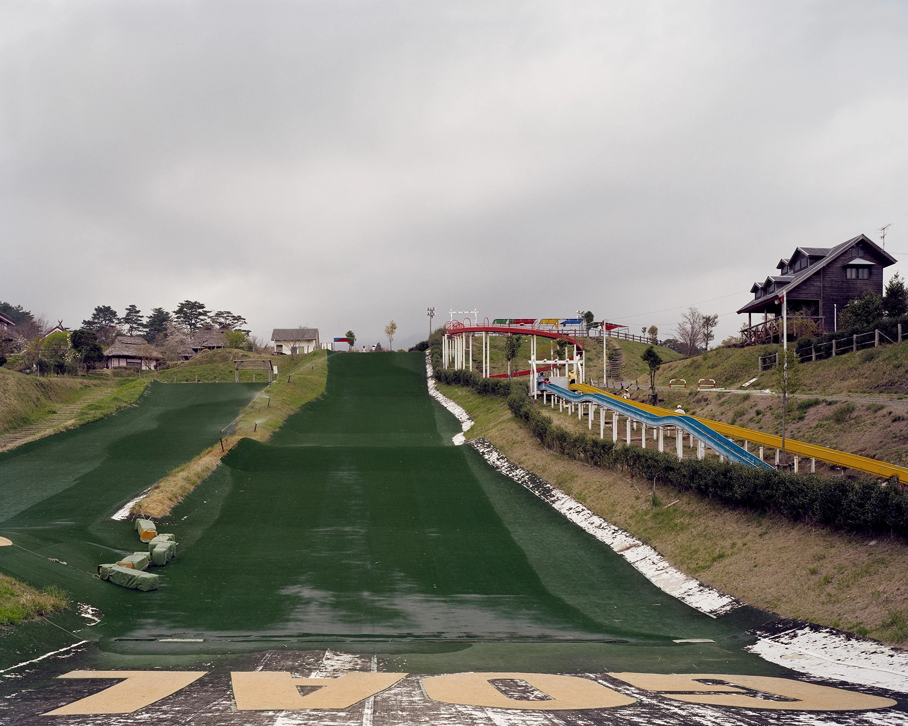 MIYAZAKI by Mark Power, photographed by Mark Power — A dry ski slope surfaced in synthetic green running up a hill, the word GOAL painted across the tarmac at its foot, a blue and yellow slide and a timber lodge on the bank to the right.