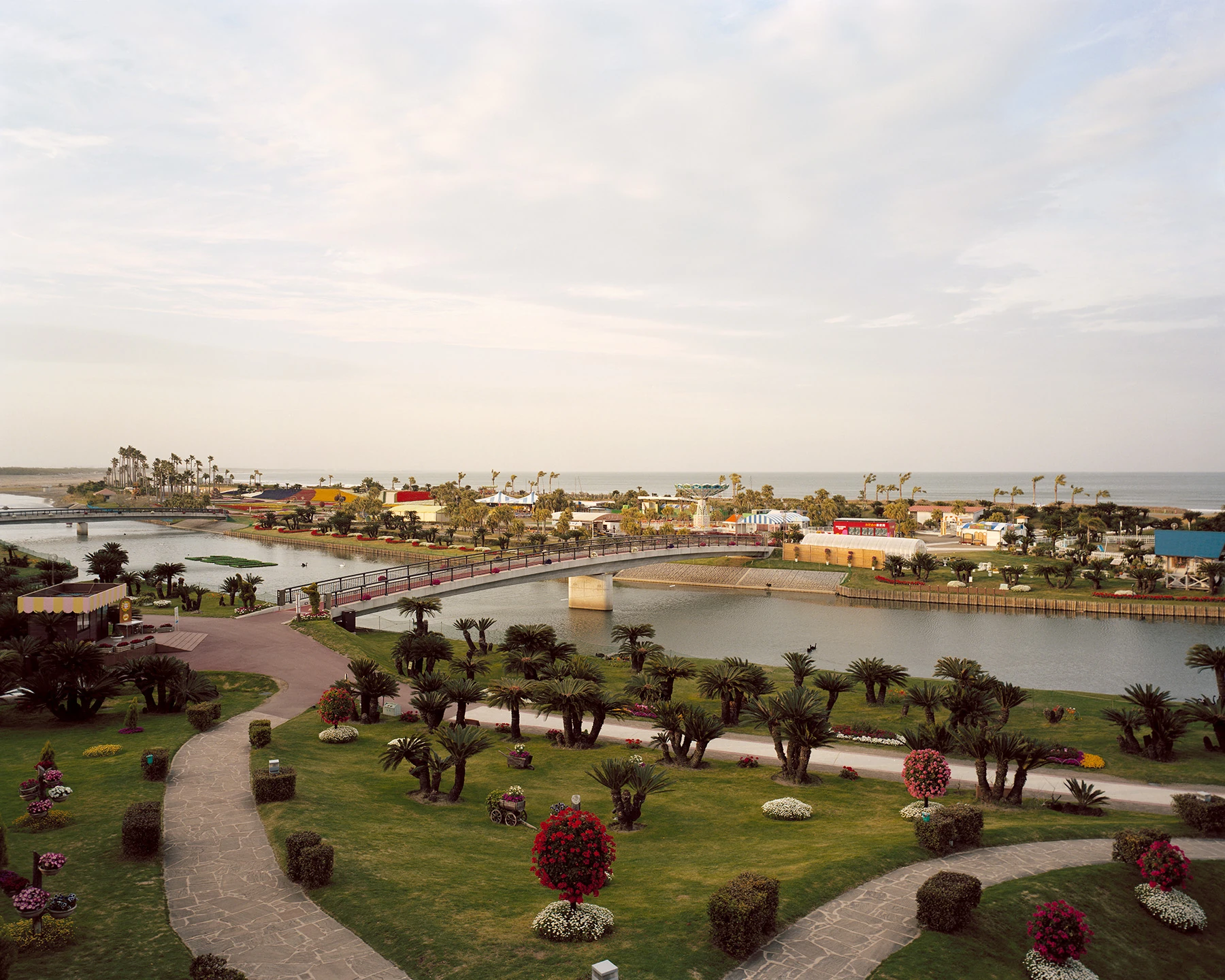 MIYAZAKI by Mark Power, photographed by Mark Power — An elevated view over a resort park, palms and flowering standards on clipped lawns, a footbridge crossing a lagoon toward fairground tents and the open sea beyond.