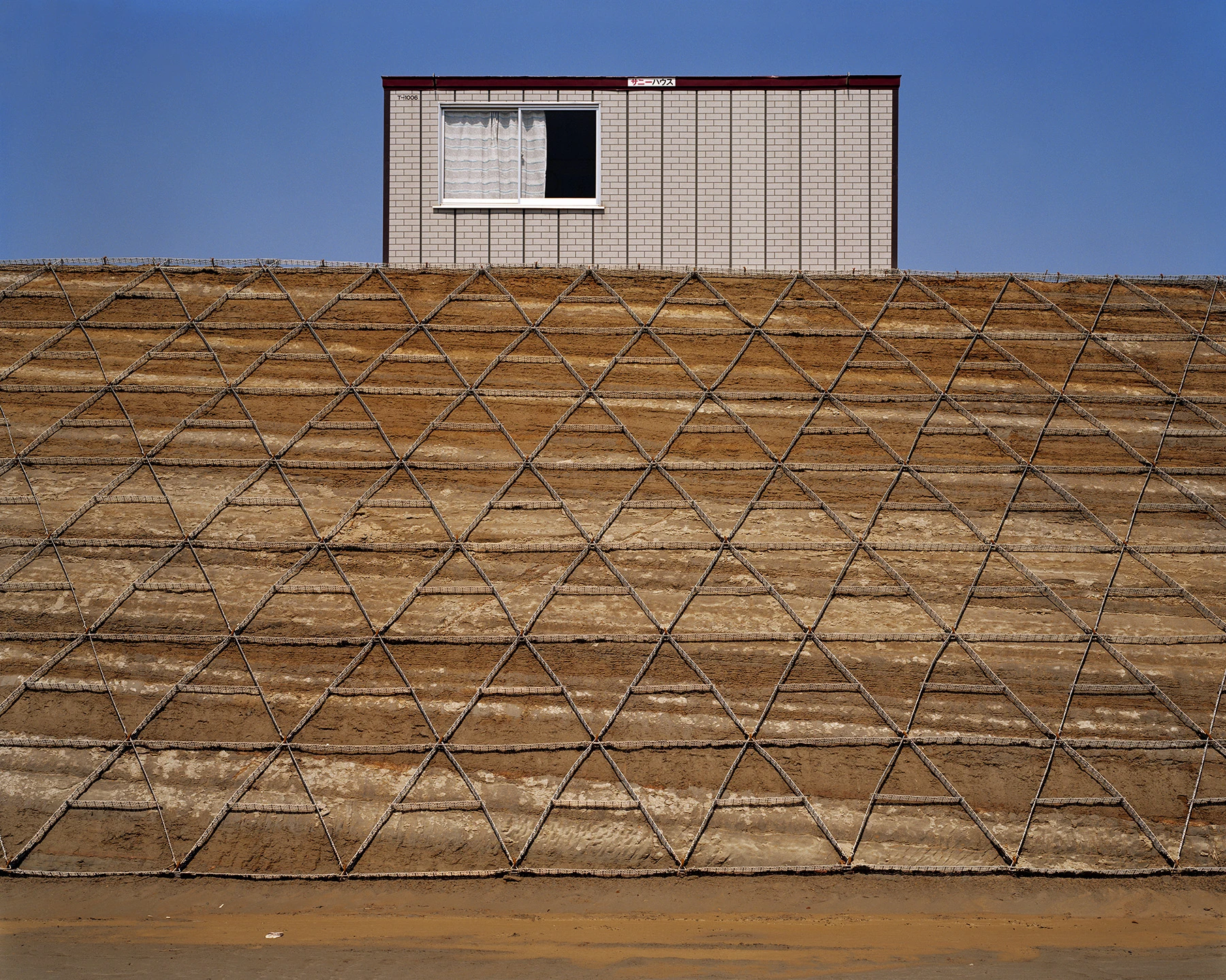 MIYAZAKI by Mark Power, photographed by Mark Power — A steep earth embankment gridded with triangular wire mesh, a grey prefabricated cabin with one curtained window standing on the ridge against a deep blue sky.