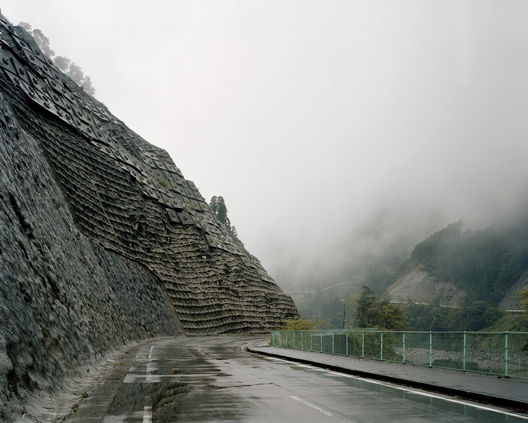 MIYAZAKI by Mark Power, photographed by Mark Power — A wet mountain road curving beneath a slope faced in overlapping black anchor plates, fog closing off the valley and a second engineered slope visible across it.