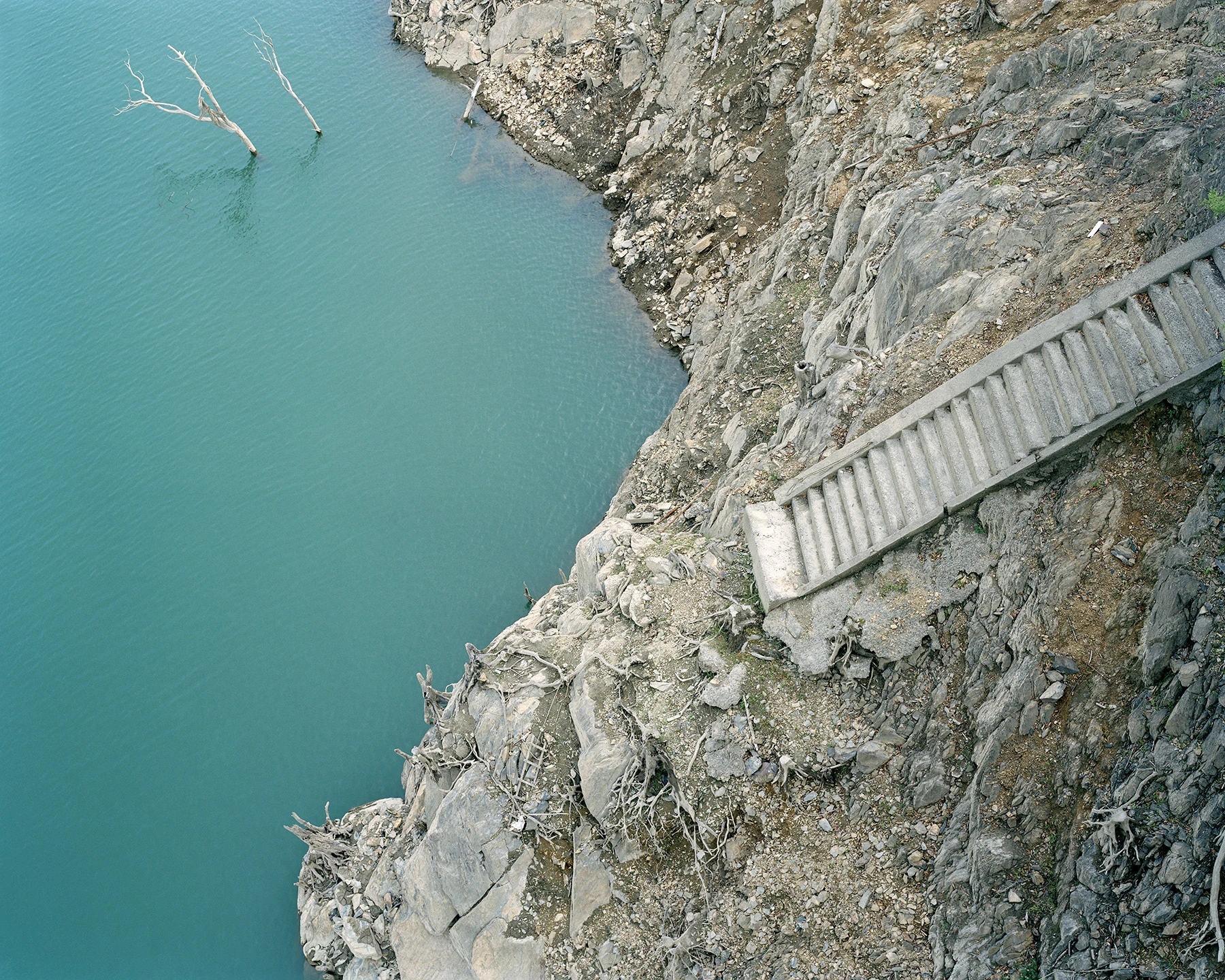 MIYAZAKI by Mark Power, photographed by Mark Power — A turquoise reservoir seen from above, bare dead branches standing out of the water, a concrete stair descending the rocky bank at right.