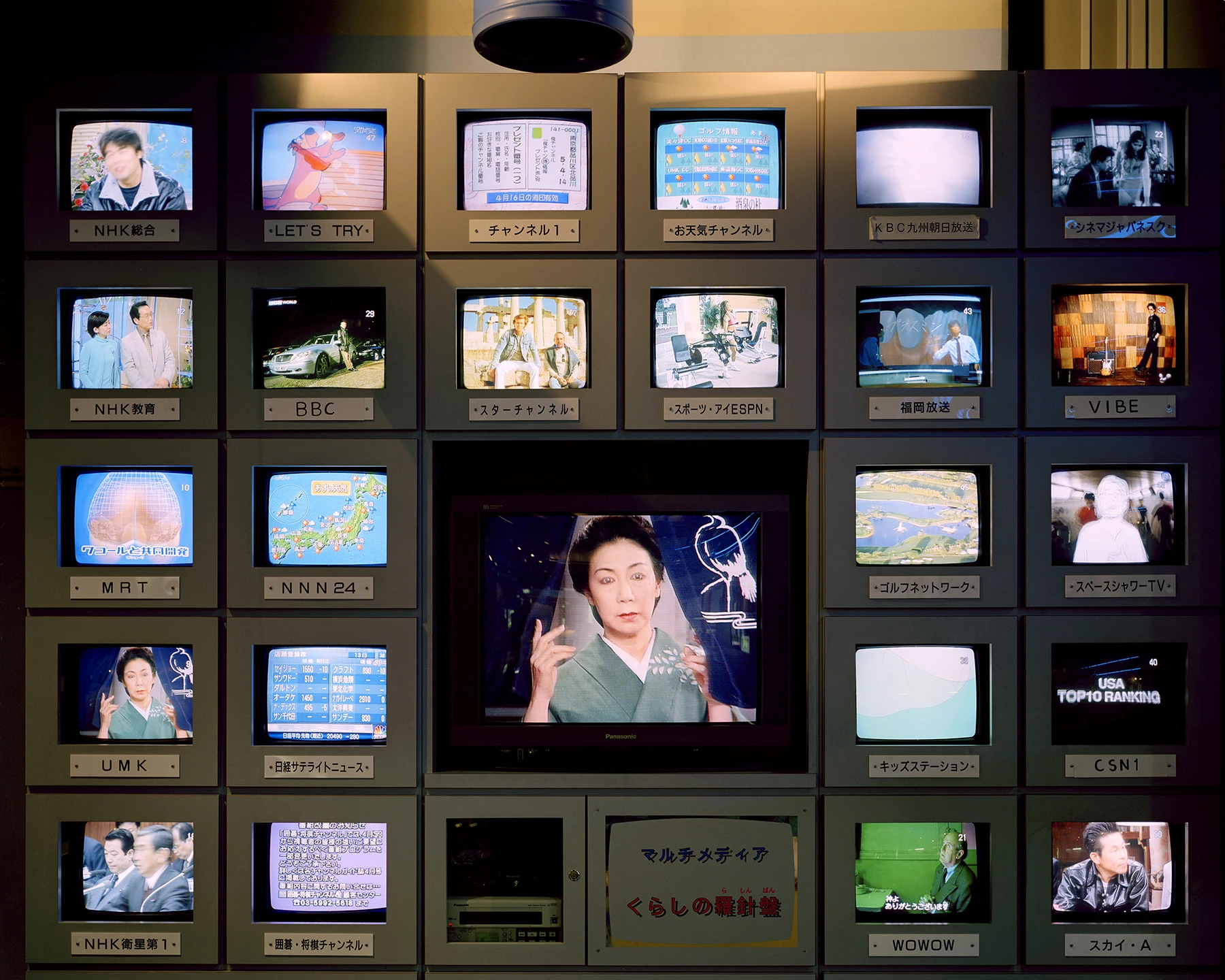 MIYAZAKI by Mark Power, photographed by Mark Power — A wall of small television monitors in labelled housings arranged around one larger central screen showing a woman in a green kimono, each set labelled with a broadcast channel.