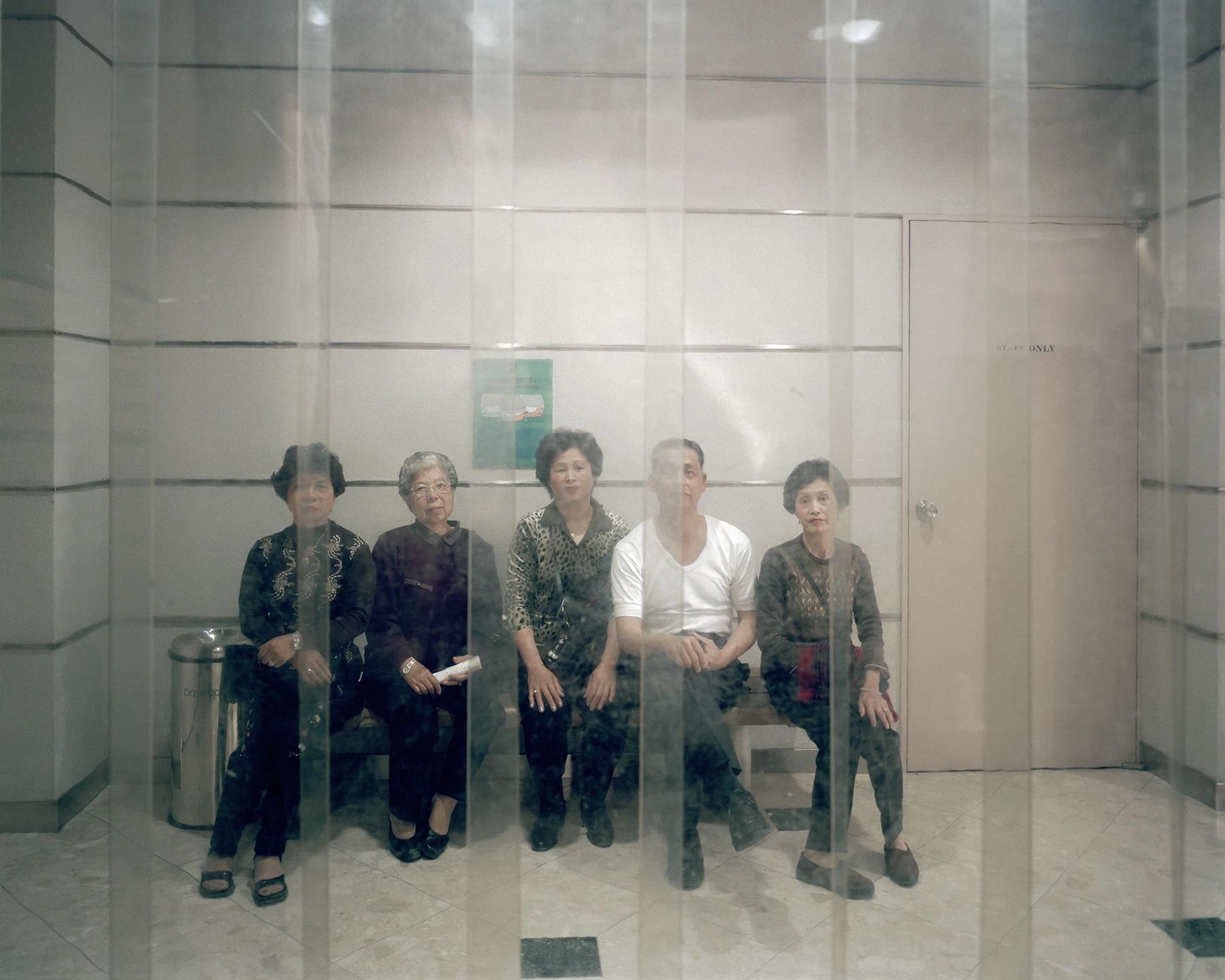 MIYAZAKI by Mark Power, photographed by Mark Power — Five older people seated on a bench in a tiled lobby, seen through a glass wall whose vertical joints throw pale bands across them, a door marked STAFF ONLY at right.