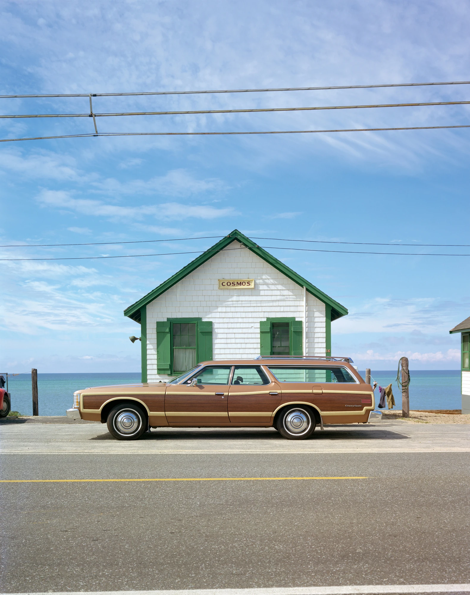 Cape Light by Joel Meyerowitz, photographed by Joel Meyerowitz — A wood-panelled Country Squire station wagon parked side-on in front of a white shingled cottage with green shutters and a COSMOS nameboard, flat blue ocean along the horizon behind.