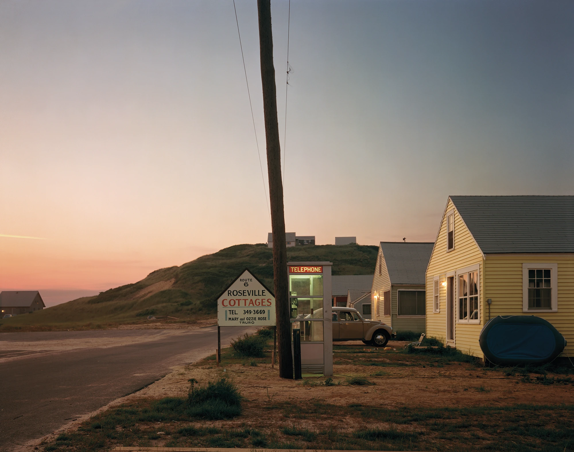 Cape Light by Joel Meyerowitz, photographed by Joel Meyerowitz — A telephone booth lit green beside a leaning utility pole at dusk, the Roseville Cottages sign at left and a row of yellow clapboard cabins to the right, the sky moving from violet to…