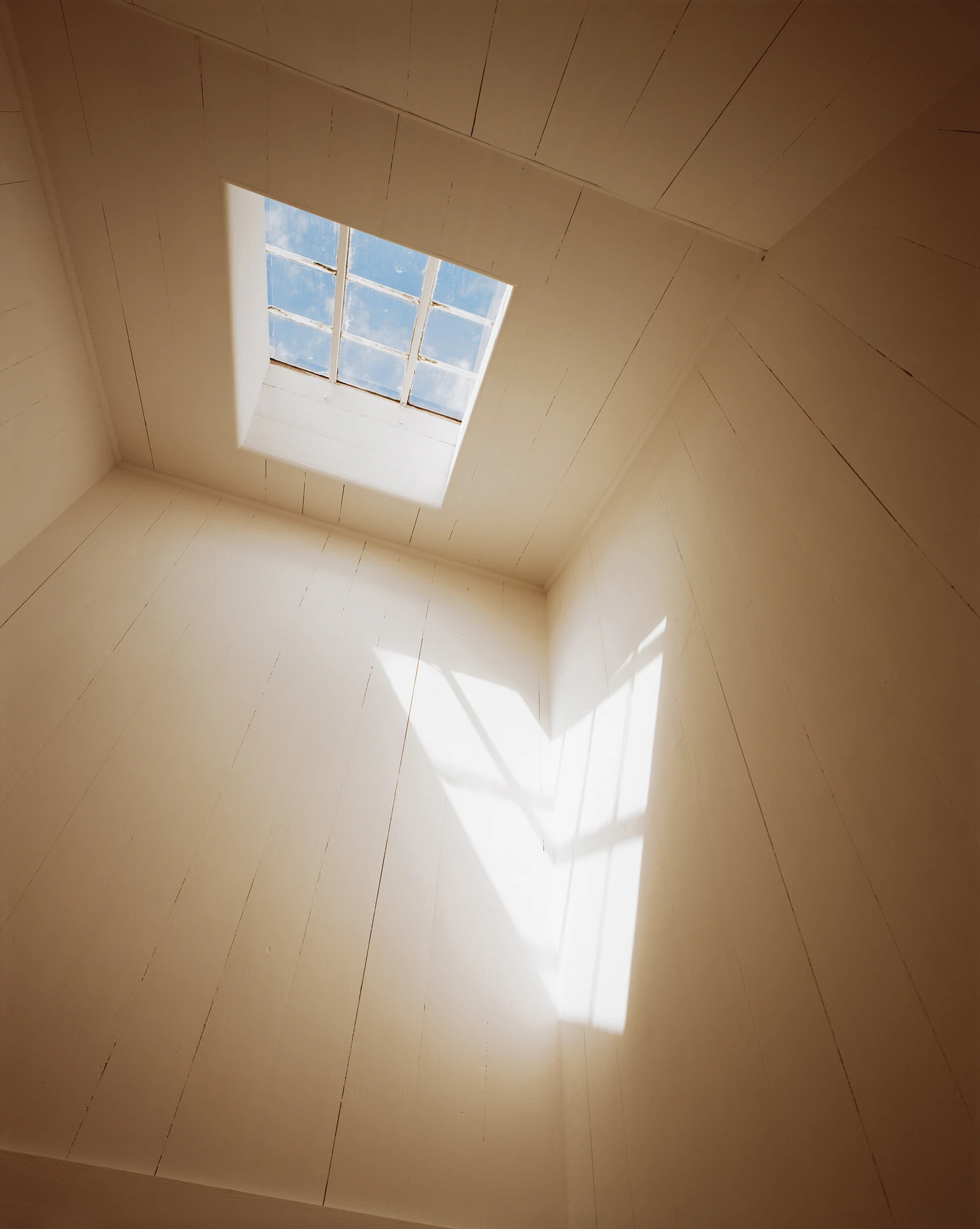 Cape Light by Joel Meyerowitz, photographed by Joel Meyerowitz — A small nine-pane skylight set into the boarded ceiling of an empty white room, a single wedge of sunlight falling across the wall and floor.