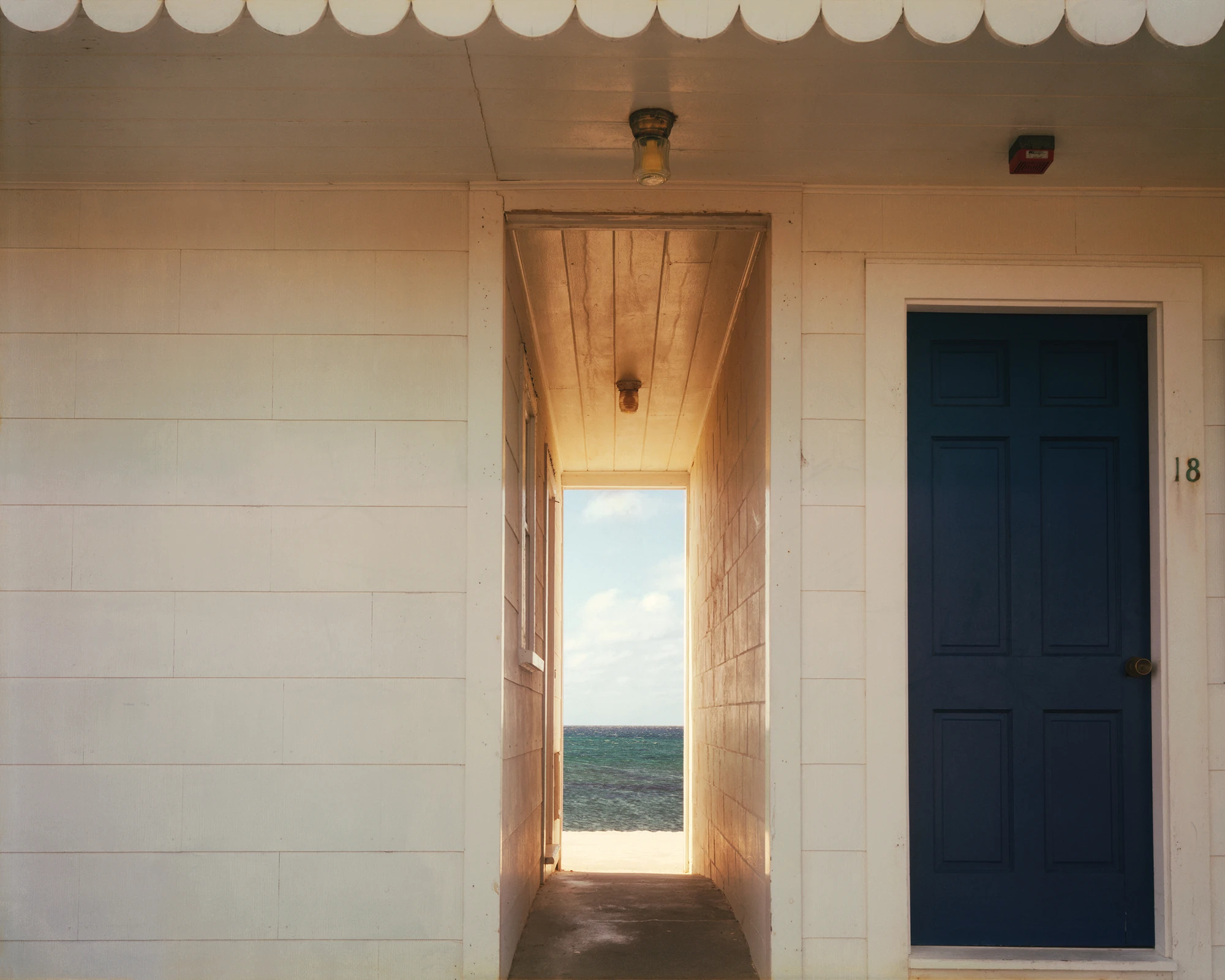 Cape Light by Joel Meyerowitz, photographed by Joel Meyerowitz — A narrow passage between two white clapboard cottages framing a bright rectangle of white sand, turquoise sea and cloud, a navy panelled door numbered 18 at right.