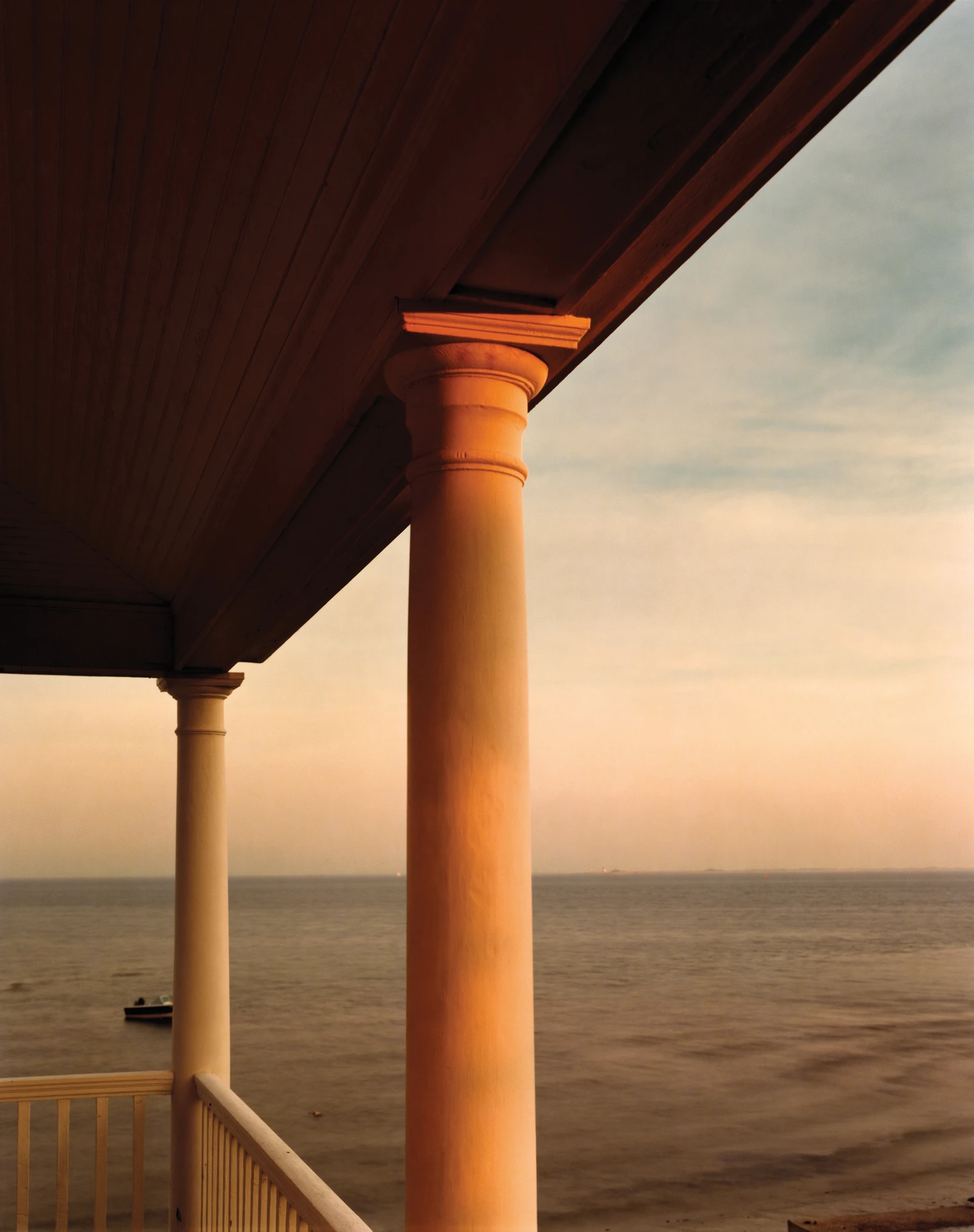 Cape Light by Joel Meyerowitz, photographed by Joel Meyerowitz — Two painted porch columns against a still bay at sunset, the nearer column washed orange by low sun, a small moored boat at the left edge.