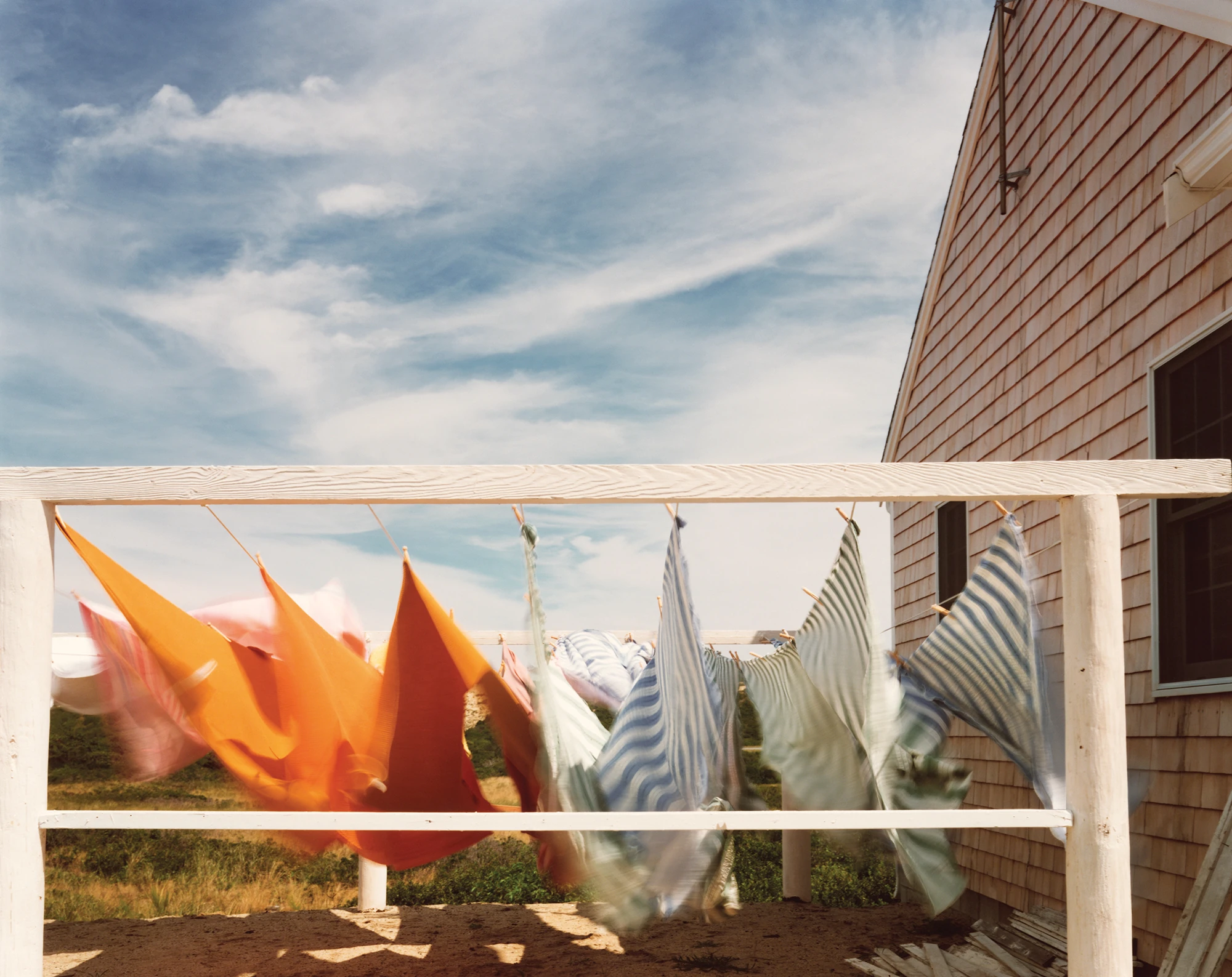 Cape Light by Joel Meyerowitz, photographed by Joel Meyerowitz — Laundry pegged along a line and blown almost horizontal, orange and pink cloth at left and blue and green striped towels at right, a shingled gable to the side under a high blue sky.