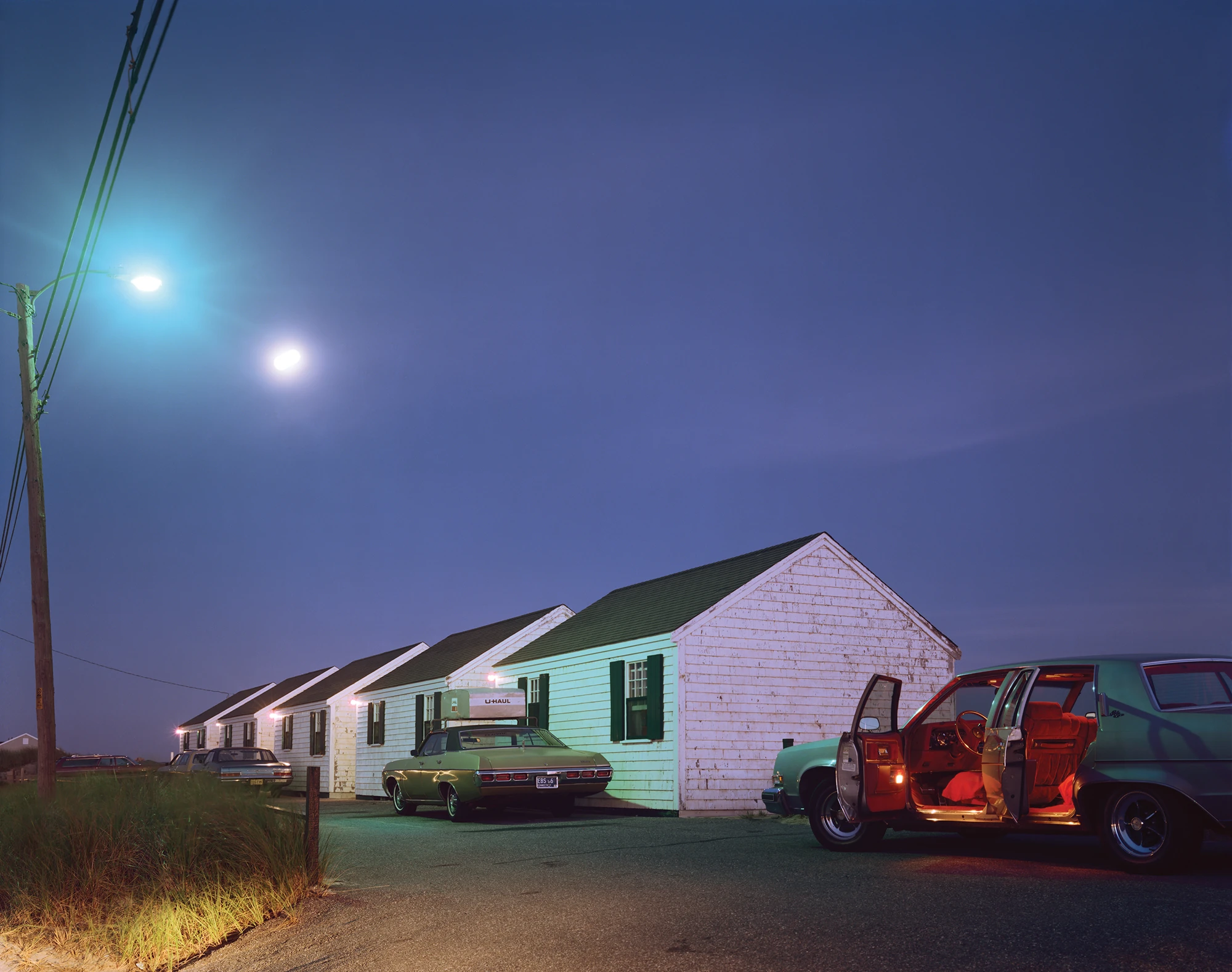 Cape Light by Joel Meyerowitz, photographed by Joel Meyerowitz — A row of cottages at night under two streetlights, a green sedan parked at the gable end and a car in the right foreground standing open with its red interior lit from within.