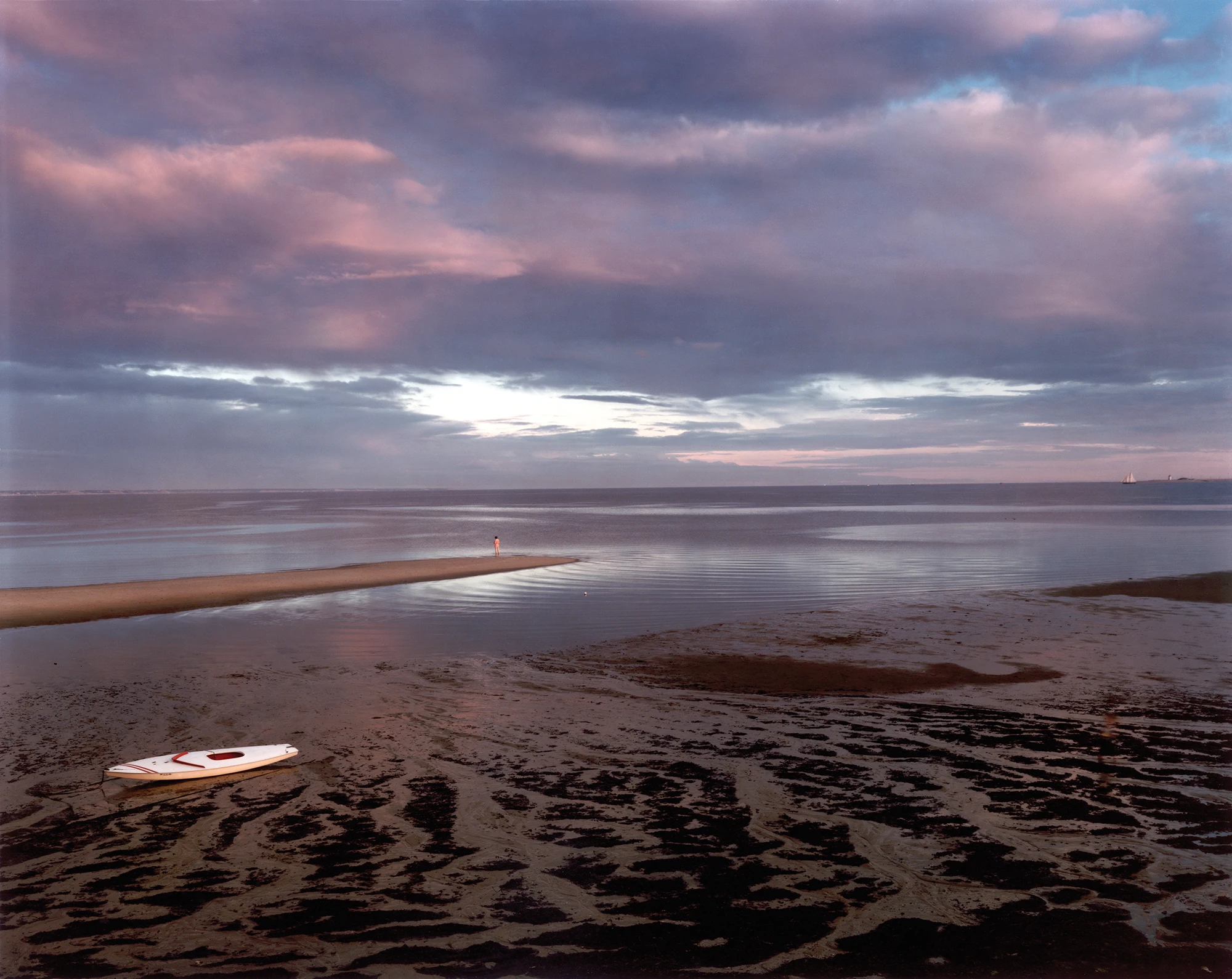 Cape Light by Joel Meyerowitz, photographed by Joel Meyerowitz — A sandbar reaching into shallow water under a mauve and pink evening sky, a lone figure standing at its far tip, a white kayak beached on the dark rippled flats in the foreground.