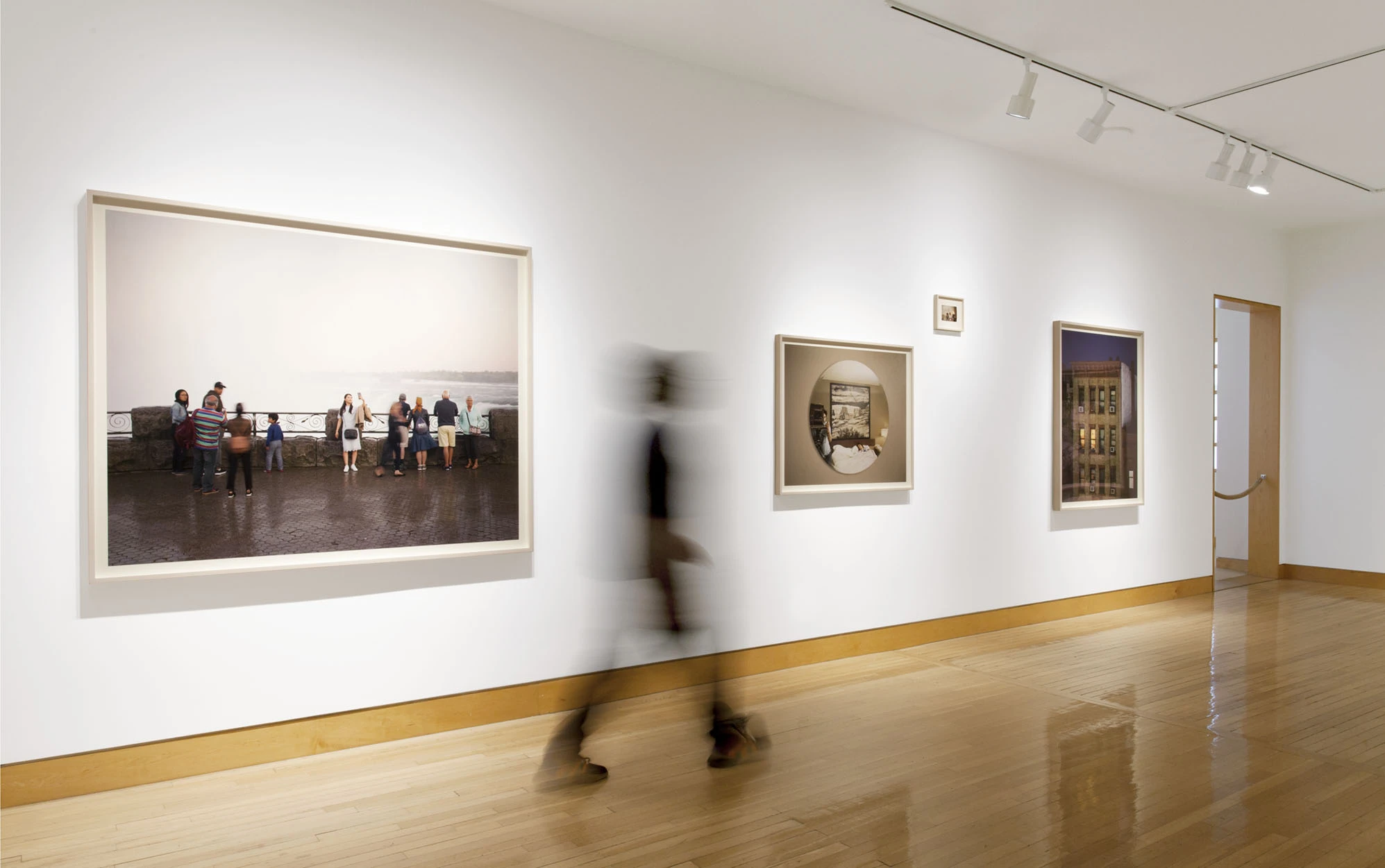 Installation view, framed prints from A Pound of Pictures hung along a white gallery wall above a pale wood floor, a visitor crossing the room in blur.