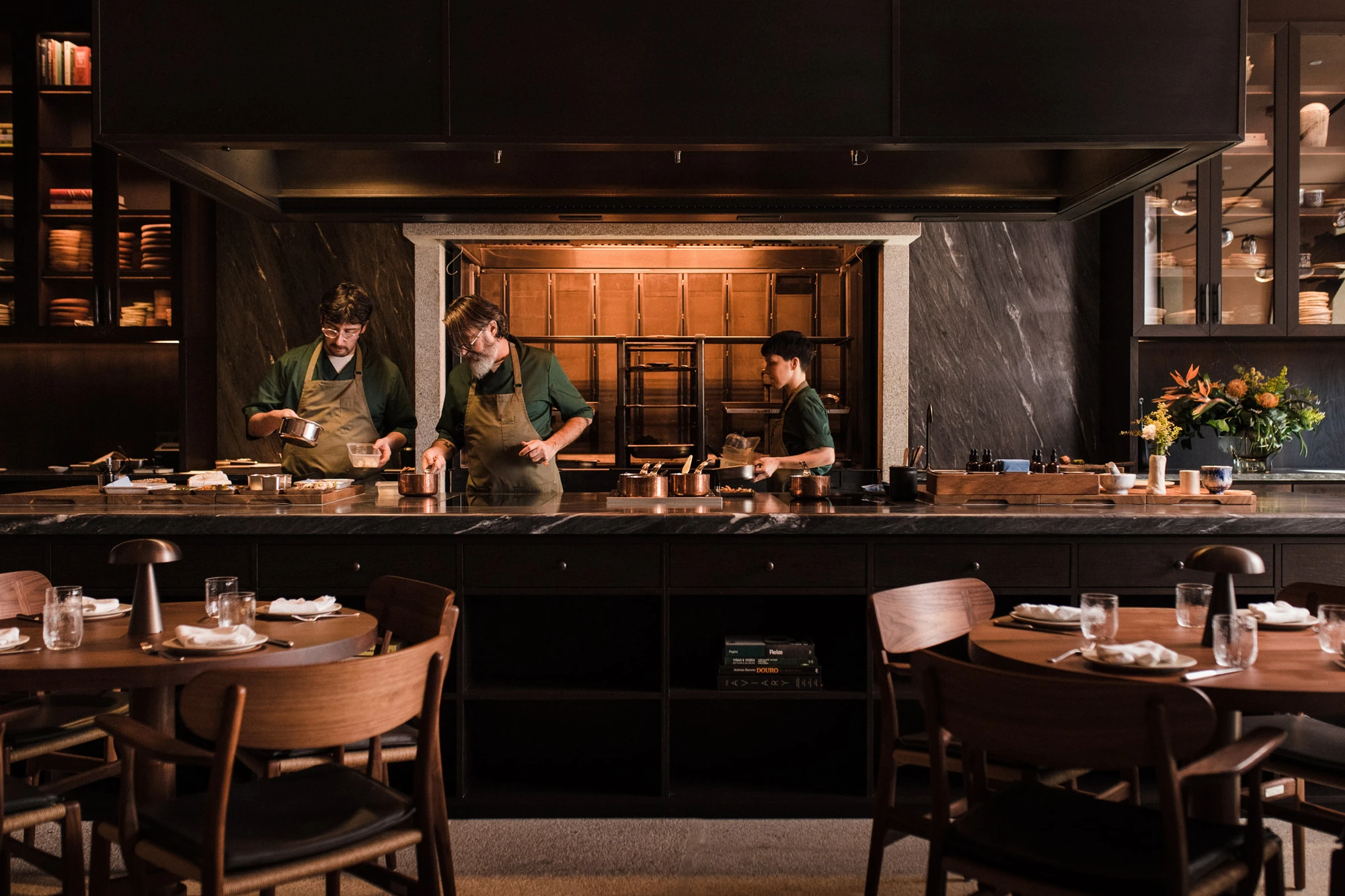 Cozinha das Flores Mural by Álvaro Siza, photographed by Luís Moreira — Cooks working along the dark grey quartzite counter of the open kitchen at Cozinha das Flores, under a black steel canopy with a brass-lined hearth behind.