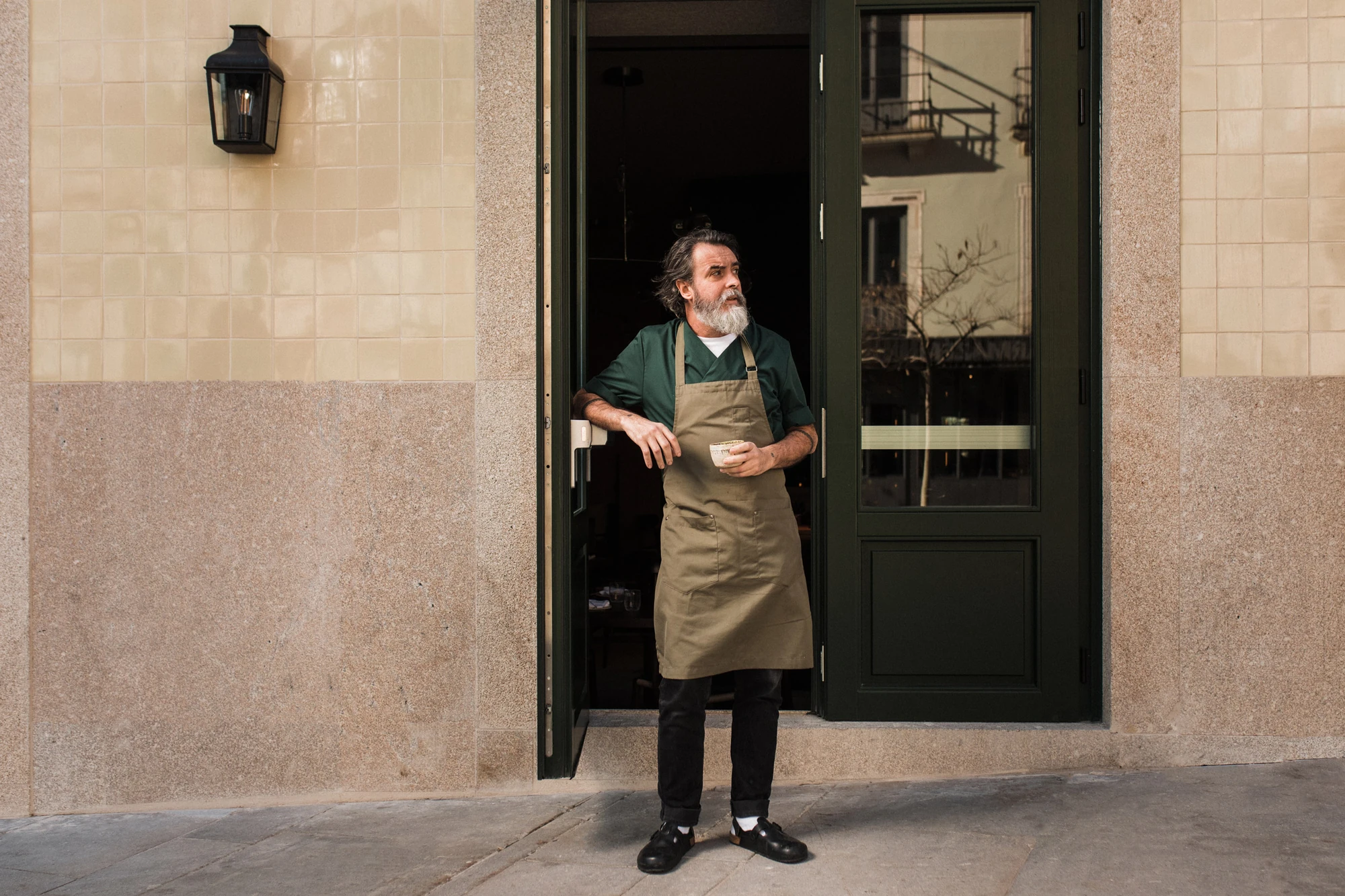 Cozinha das Flores Mural by Álvaro Siza, photographed by Luís Moreira — A chef in a green jacket and canvas apron standing in the dark green doorway of Cozinha das Flores, between cream tile and speckled granite on Largo de São Domingos.
